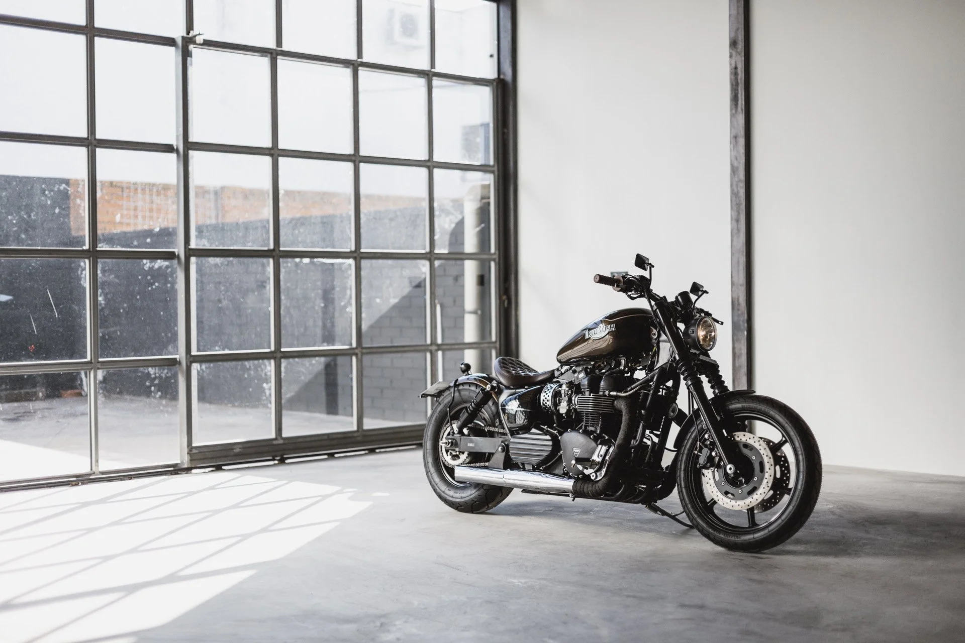 Motocicleta Triumph bobber en un espacio con paredes blancas y un gran ventanal de vidrio que deja entrar luz natural.