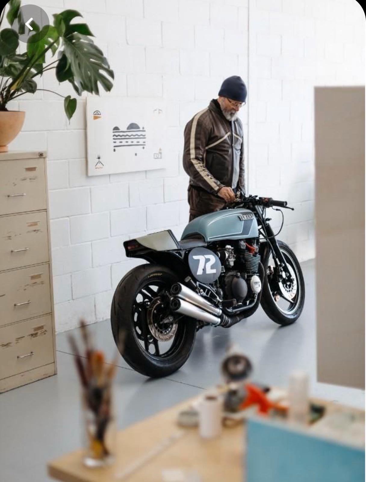 Un hombre con gorro y chaqueta de motociclismo junto a una motocicleta clásica con el número 72, en un ambiente de exposición de arte.