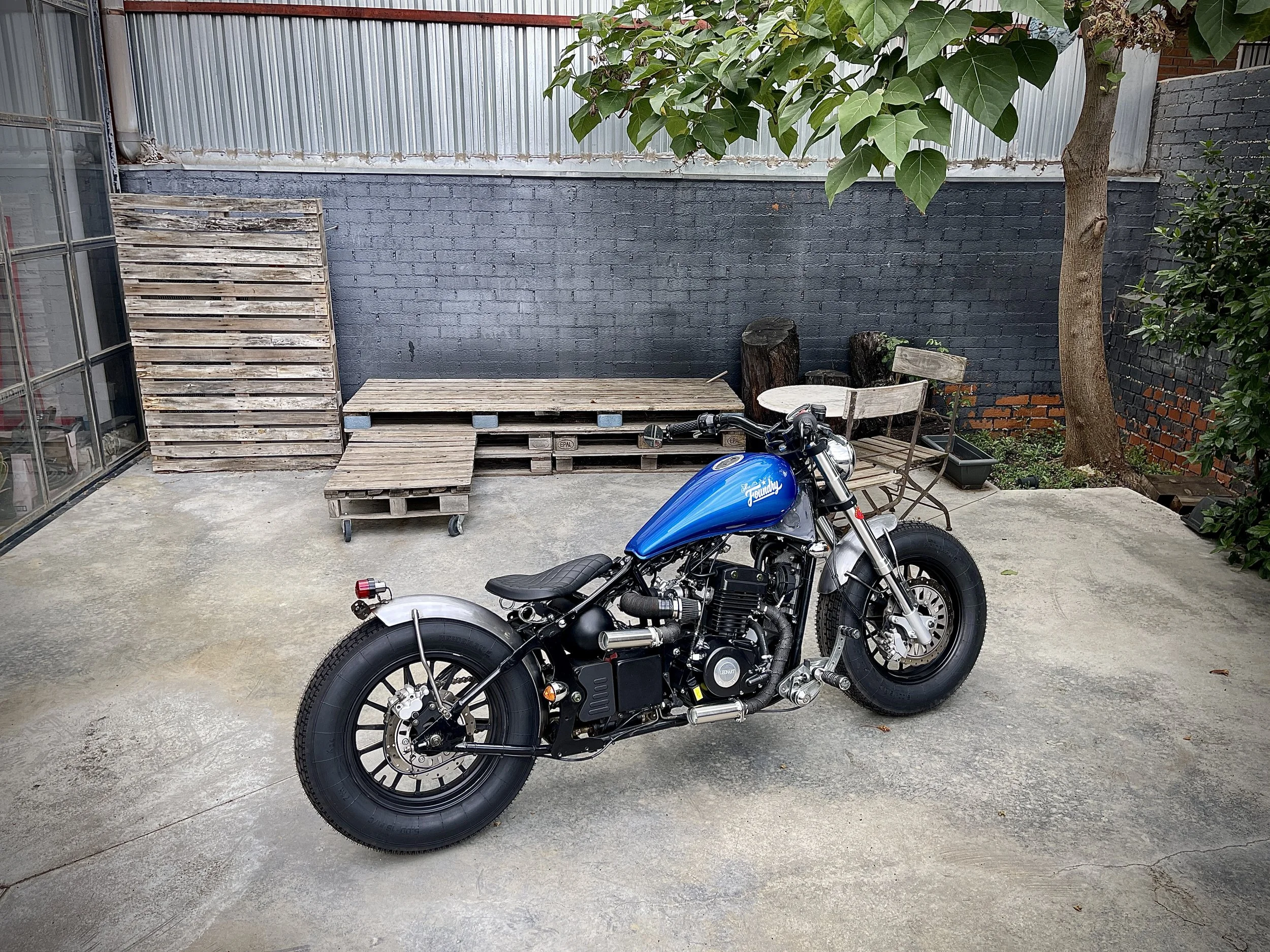 Una motocicleta bobber estacionada en un patio con un muro de ladrillos pintado en gris y una cerca de lámina metálica en el fondo, junto a una mesa y sillones hechos con palets de madera, y un árbol con hojas grandes a la derecha.