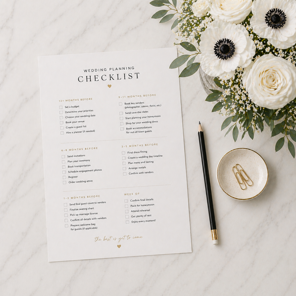 Wedding planning checklist on a white marble surface, with a black pen, a gold-accented bowl with paper clips, and a bouquet of white flowers and greenery.