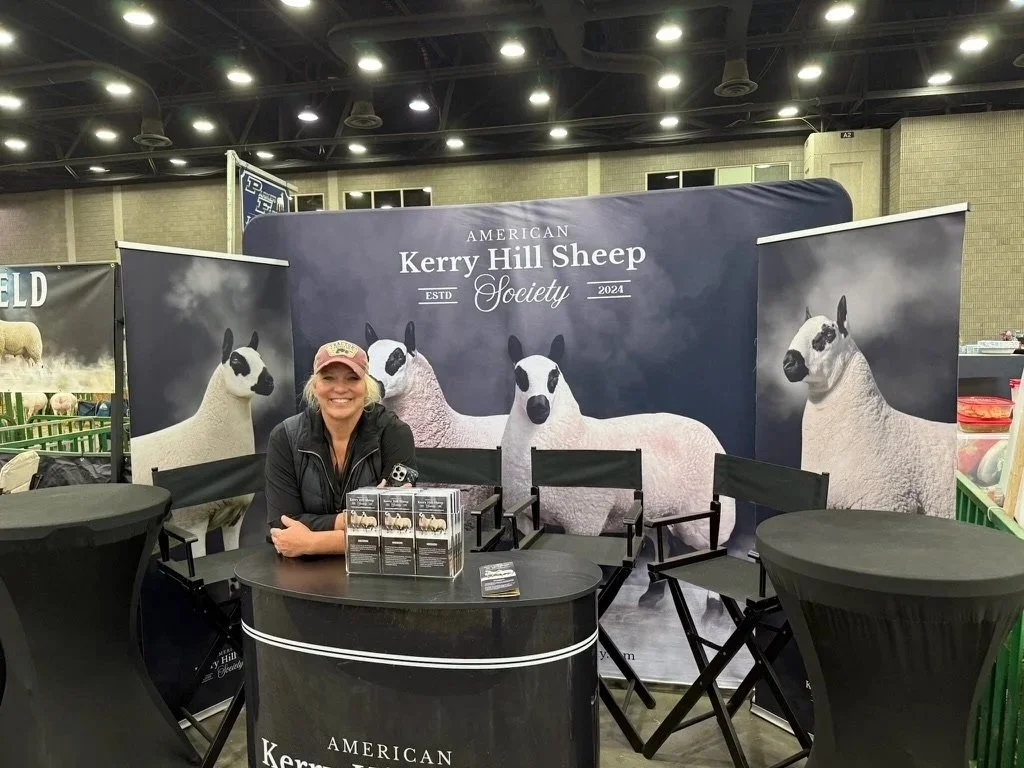 Tracy Bloom at the NAILE exhibition booth for the American Kerry Hill Sheep Society, with large banners showing black and white sheep behind her, and a table with promotional materials.