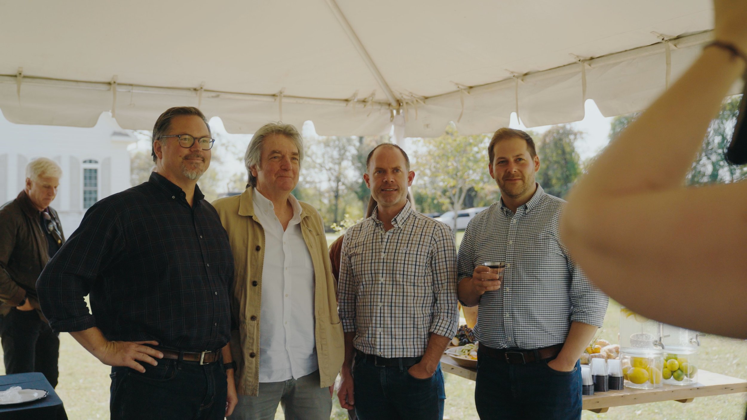 Group of five men standing under a tent at an outdoor event, with food and drinks on a table behind them.
