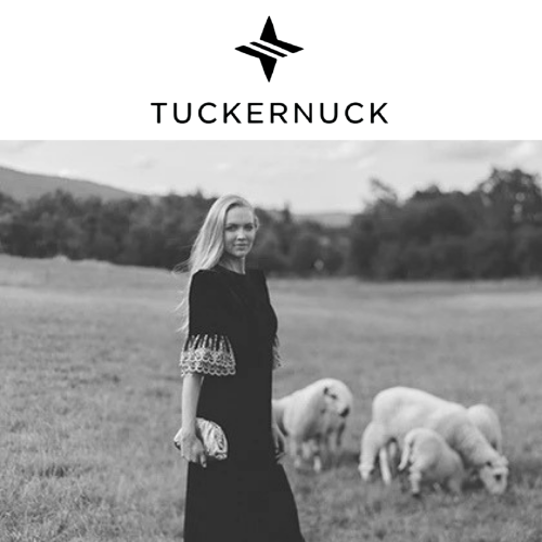 A woman standing in a field with sheep grazing behind her, and mountains in the distance. Tuckernuck
