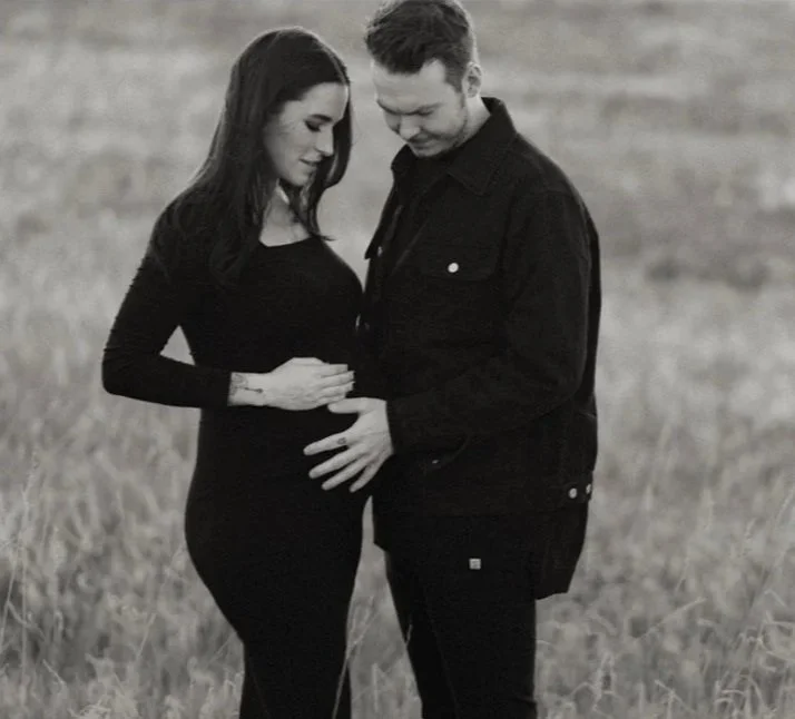 A couple stands in a field, with the woman pregnant, touching her belly and looking at each other.