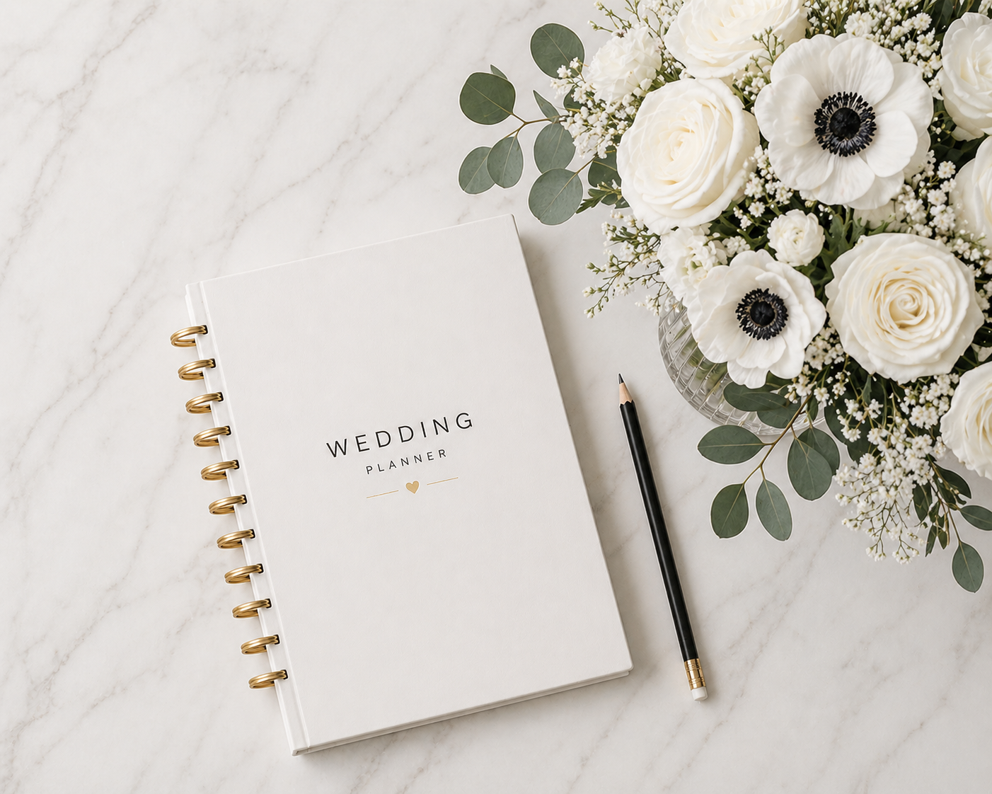 A wedding planner book, a black pen, and a flower arrangement with white roses, anemones, and greenery on a white marble surface.