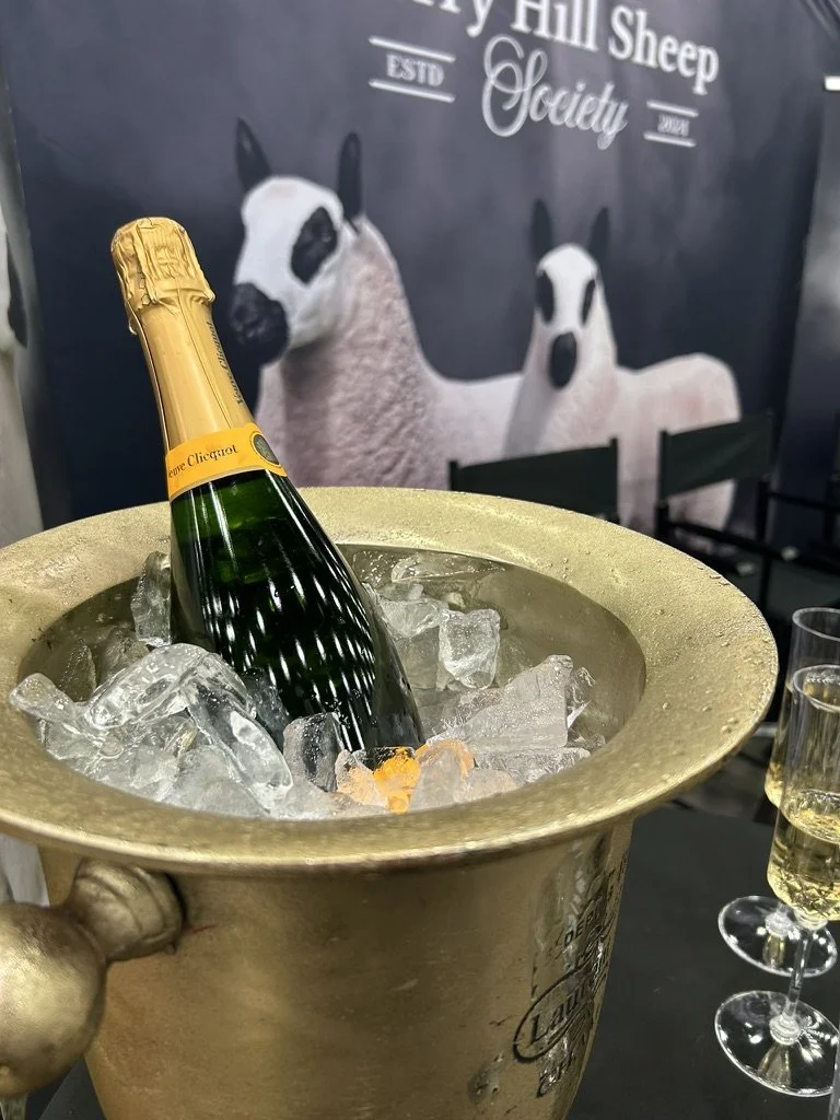 A bottle of Veuve Clicquot champagne chilling in a large gold ice bucket filled with ice cubes, with two glasses of champagne nearby. In the background, a wall with a sign reading 'American Kerry Hill Sheep Society' and an illustration of two sheep.