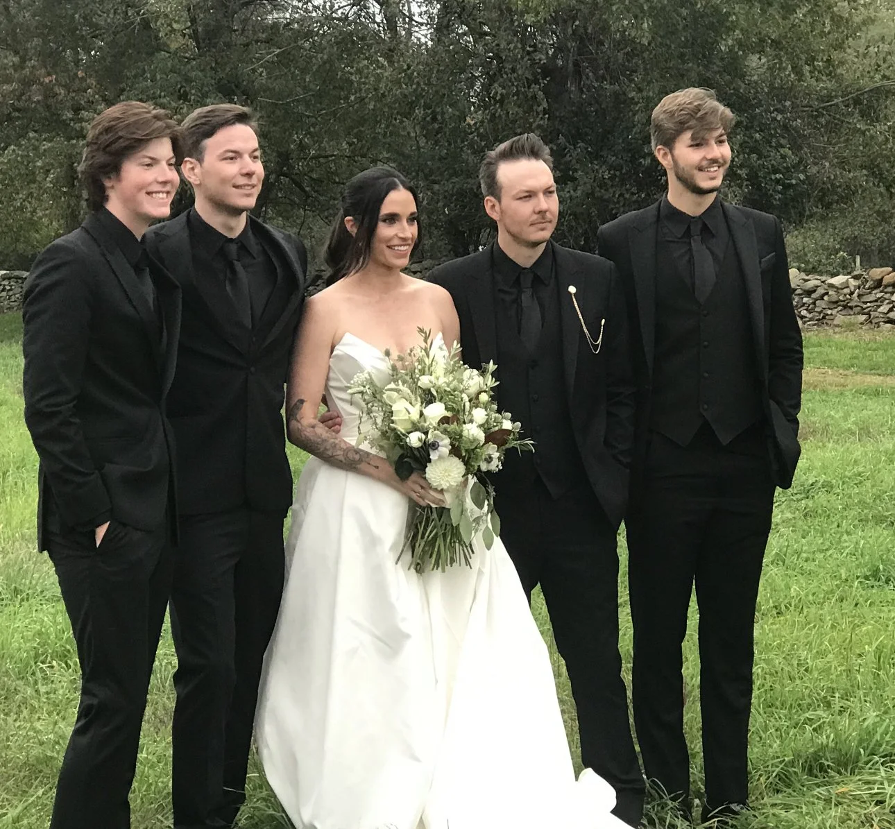 A bride in a white wedding dress holding a bouquet of white flowers standing with five men in black suits outdoors during daytime.