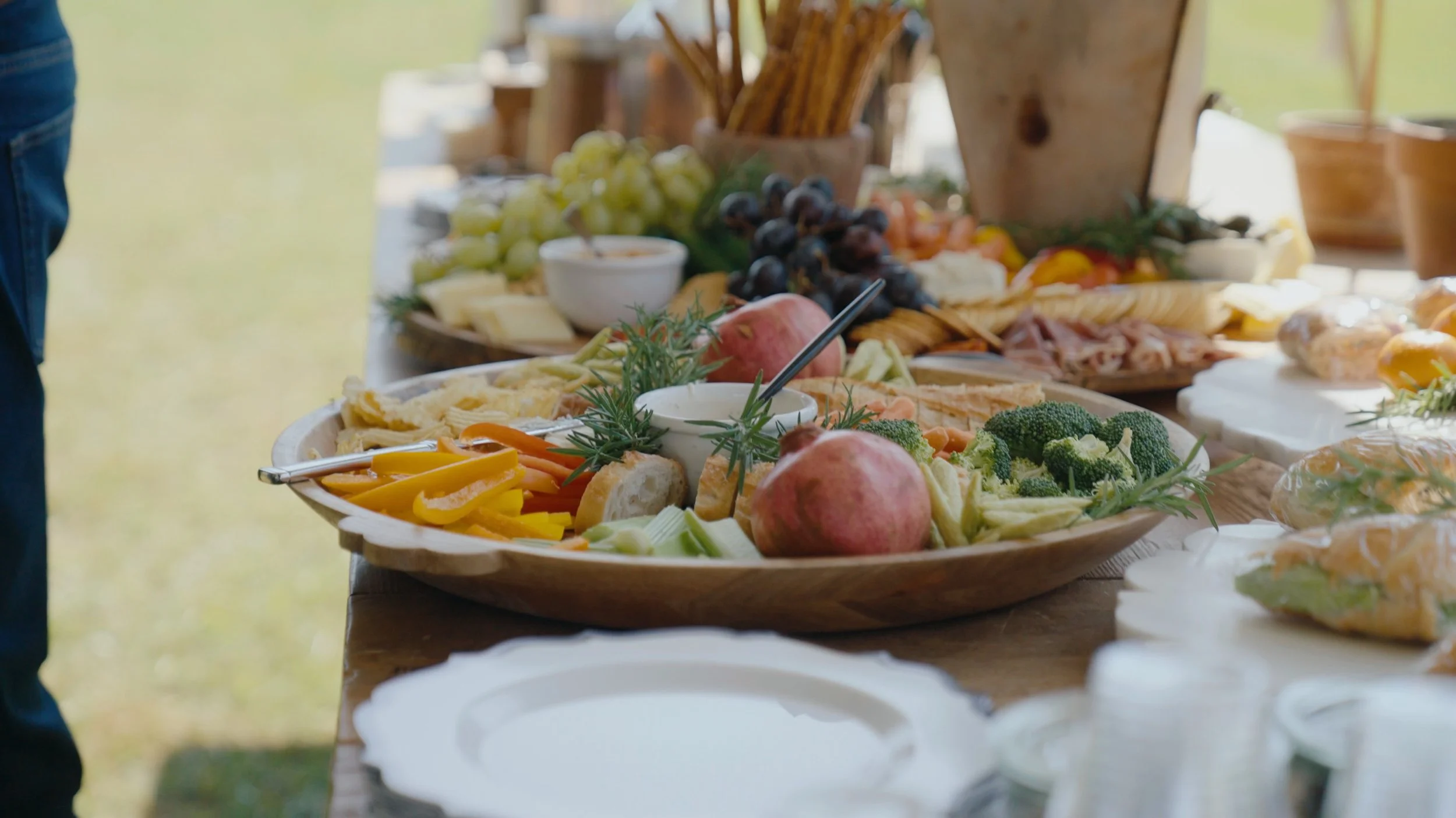 A table loaded with a wooden platter of fresh vegetables, fruits, cheeses, and bread, with grapes, pomegranate, broccoli, and sliced vegetables, set outdoors during daytime.