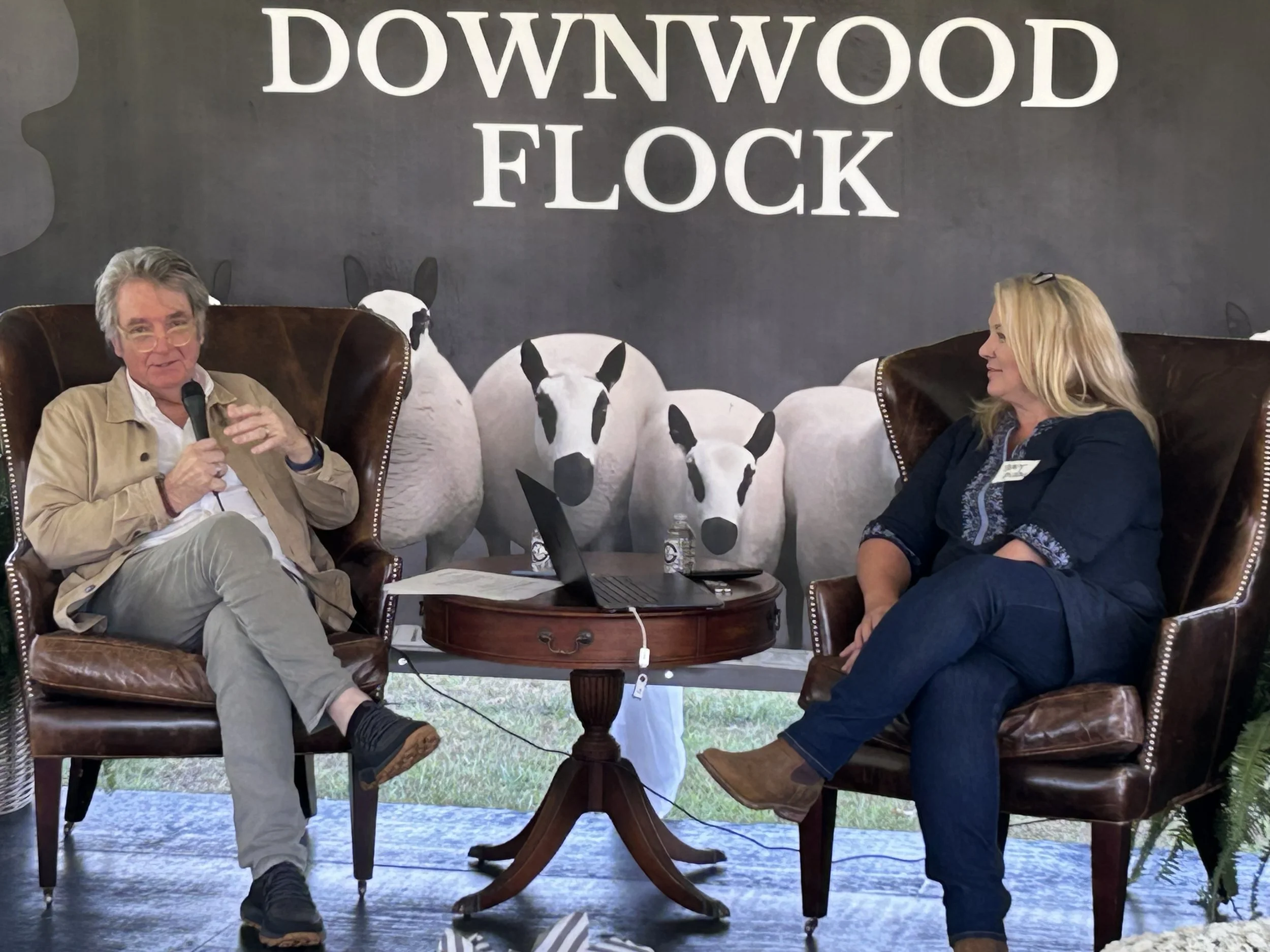 Two people sitting in leather armchairs talking at the Downwood FLOCK event, with a backdrop featuring a flock of sheep and large white text reading 'Downwood FLOCK'.