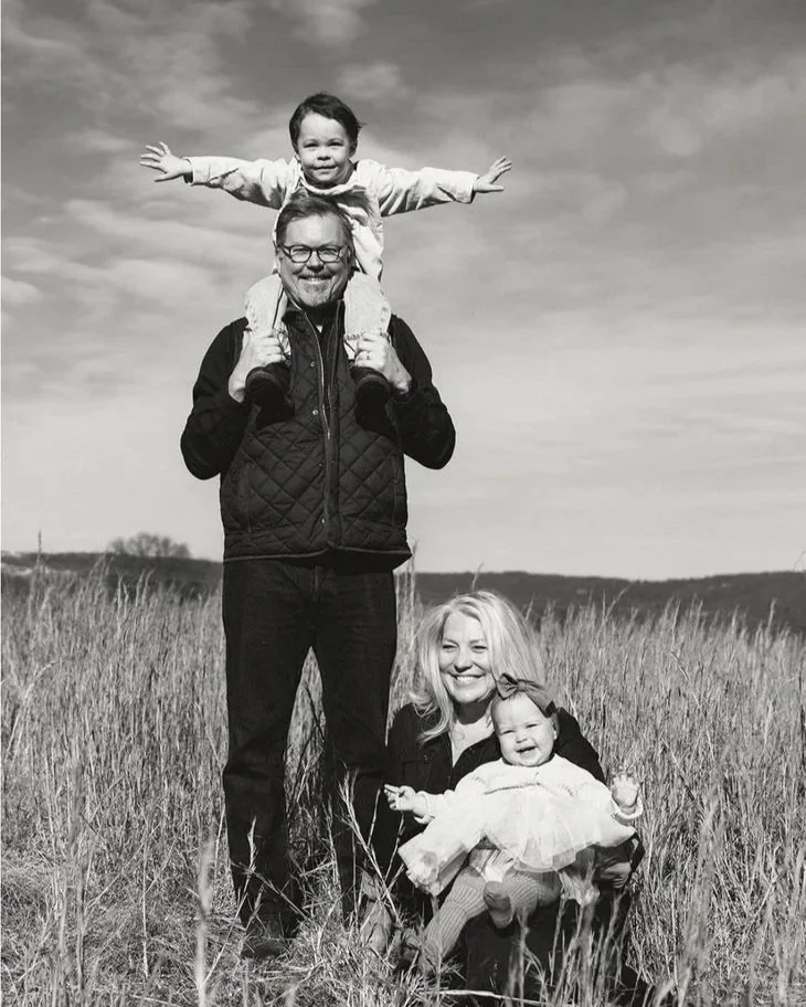 A black-and-white photo of a smiling family of four outdoors in a grassy field, with clear skies. The father, wearing glasses and a quilted vest, stands holding his daughter on his shoulders. The daughter, with arms outstretched, is smiling. The mother, kneeling beside him, holds their younger child, who is also smiling and dressed in a dress and tights.
