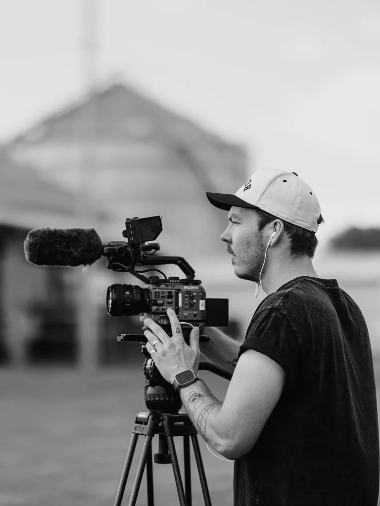 A man operating a Sony professional video camera outdoors, wearing a watch and earbuds, with a blurred building in the background.
