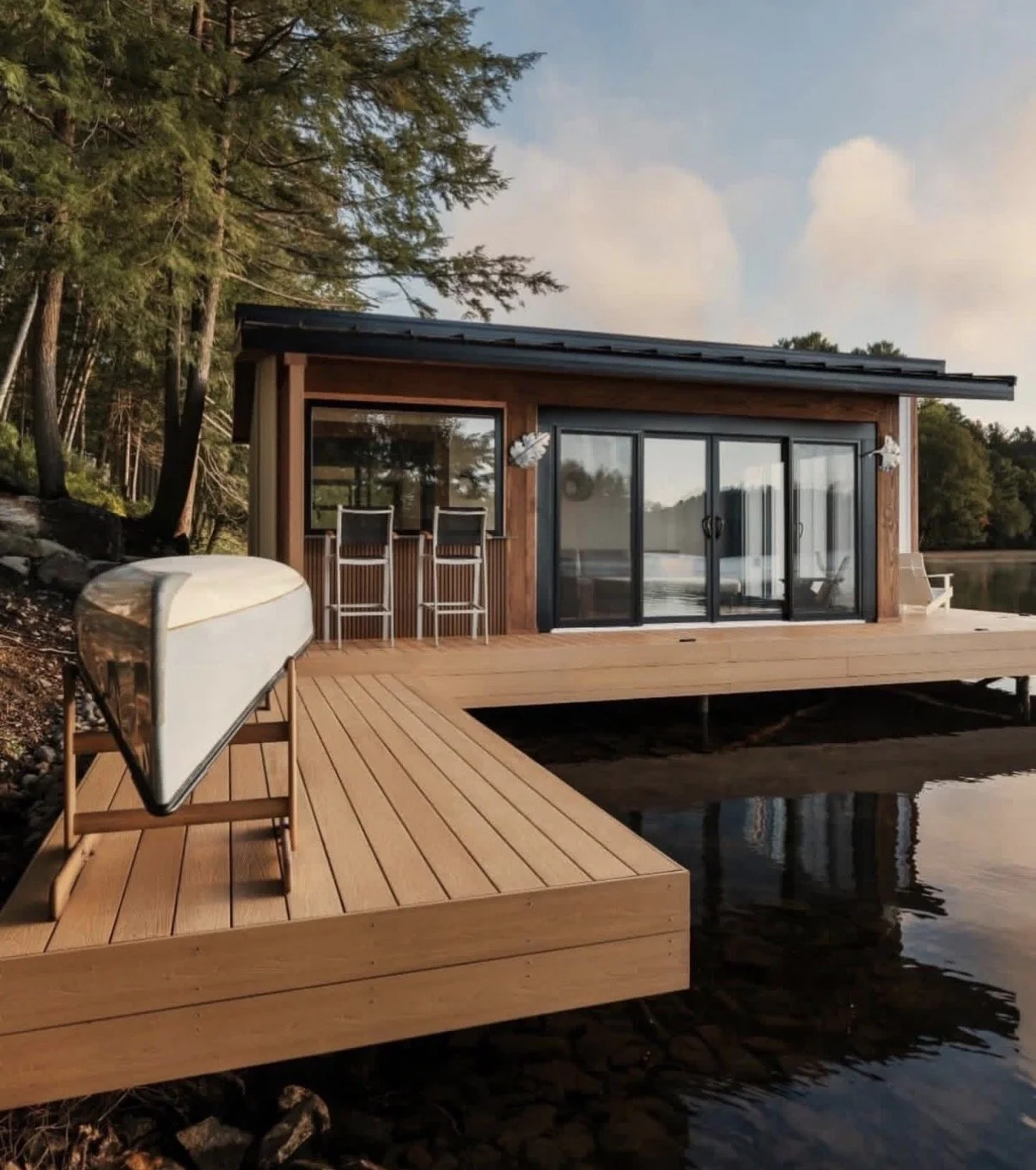 A modern lakeside cabin with a wooden deck extending over the water. The cabin features large glass sliding doors and a small outdoor seating area with two high chairs. A canoe is placed on the deck near the shore, and there are trees in the background under a partly cloudy sky.
