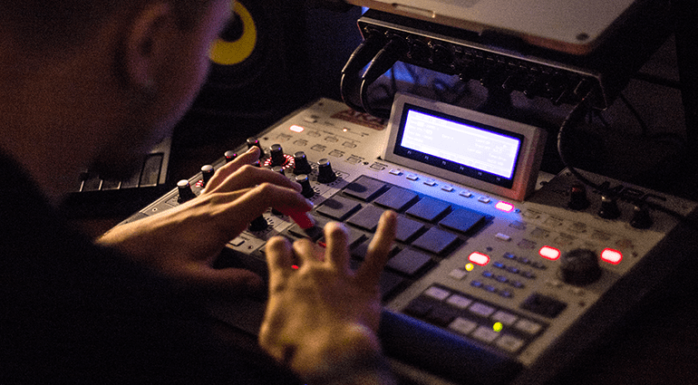 A person using a music production device with multiple pads, knobs, and buttons, in a dark studio setting.