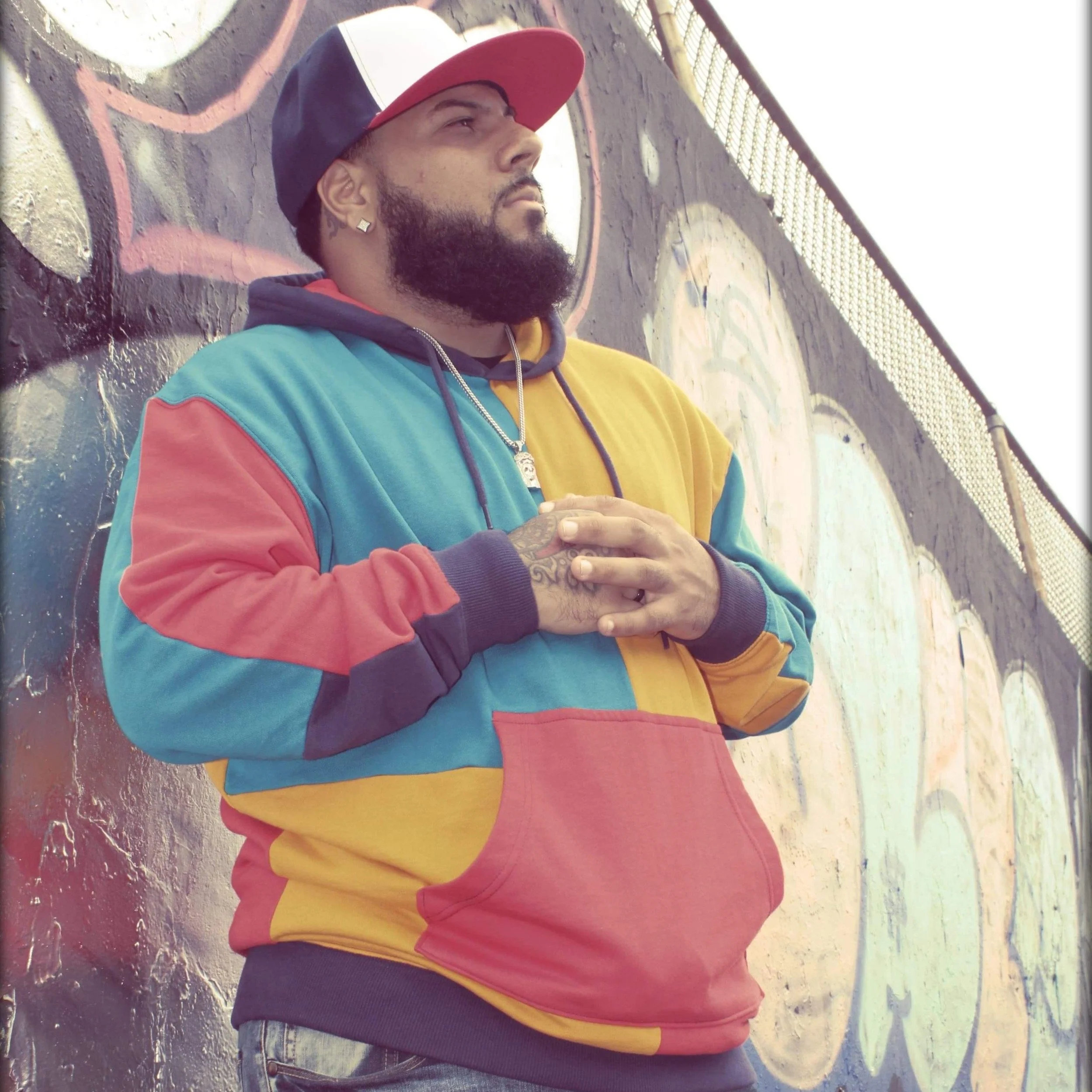 A man with a beard, earrings, wearing a multicolored hoodie and a red and gray baseball cap, leaning against a graffiti-covered wall.