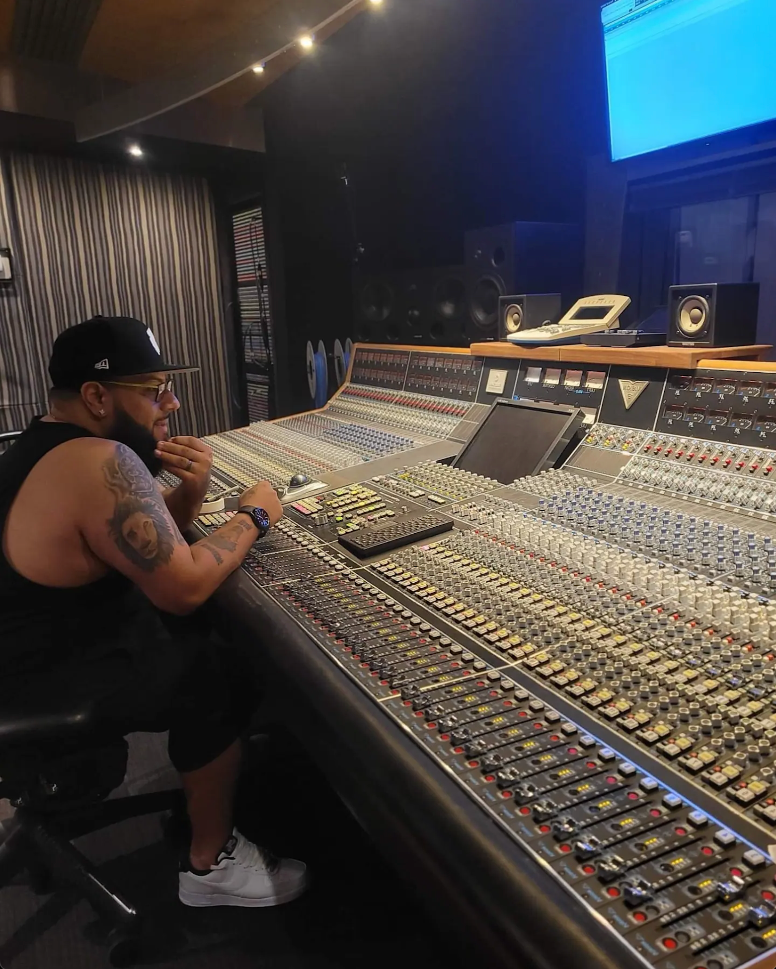 A man with tattoos, glasses, a black cap, and white sneakers sitting in front of a large mixing console in a recording studio, with multiple screens, speakers, and audio equipment around him.
