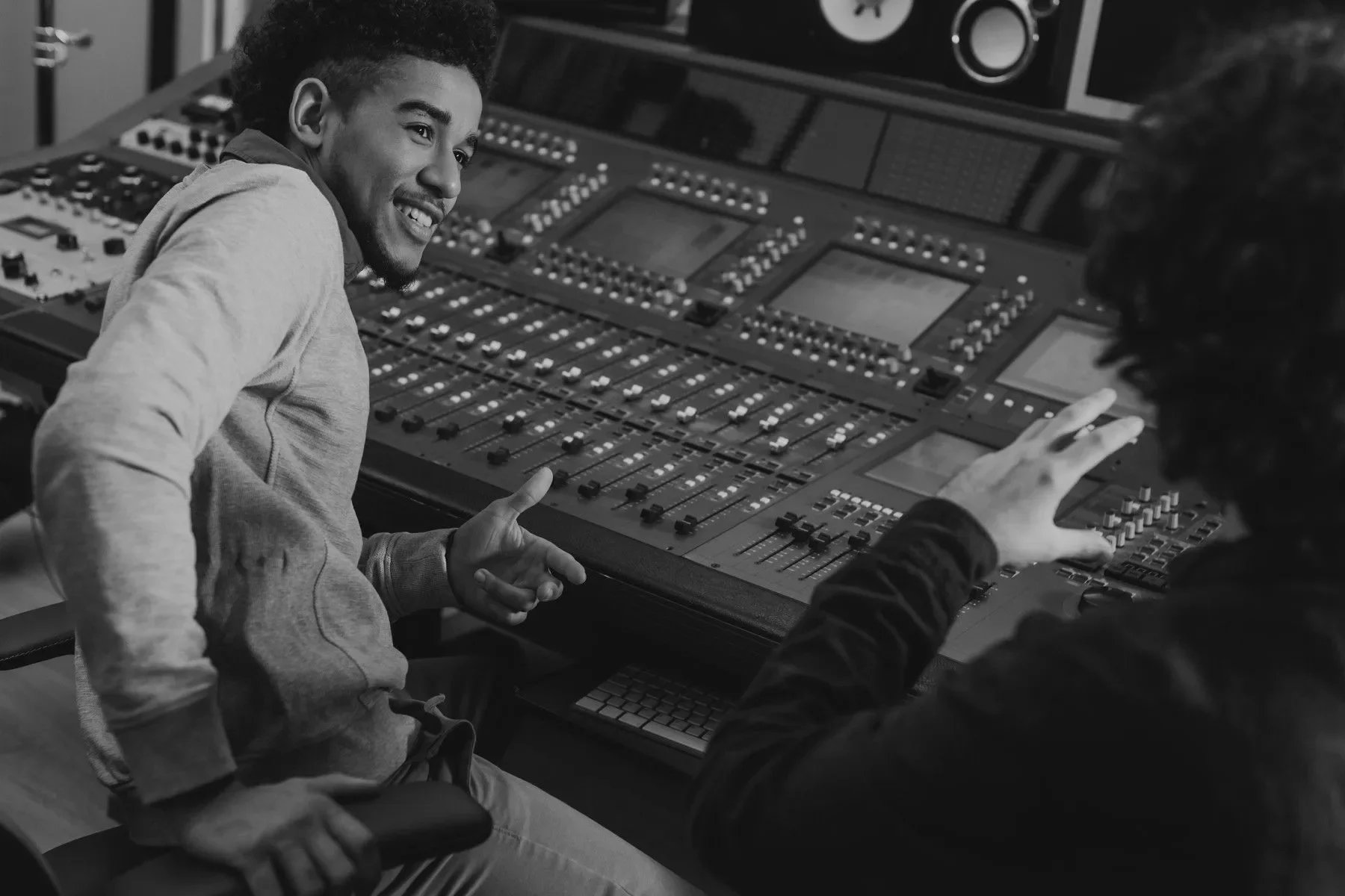 Two young men are in a recording studio, sitting at a mixing console. One man is smiling and gesturing with his hands, while the other man's hand is on the console. The studio has large speakers mounted on the wall.