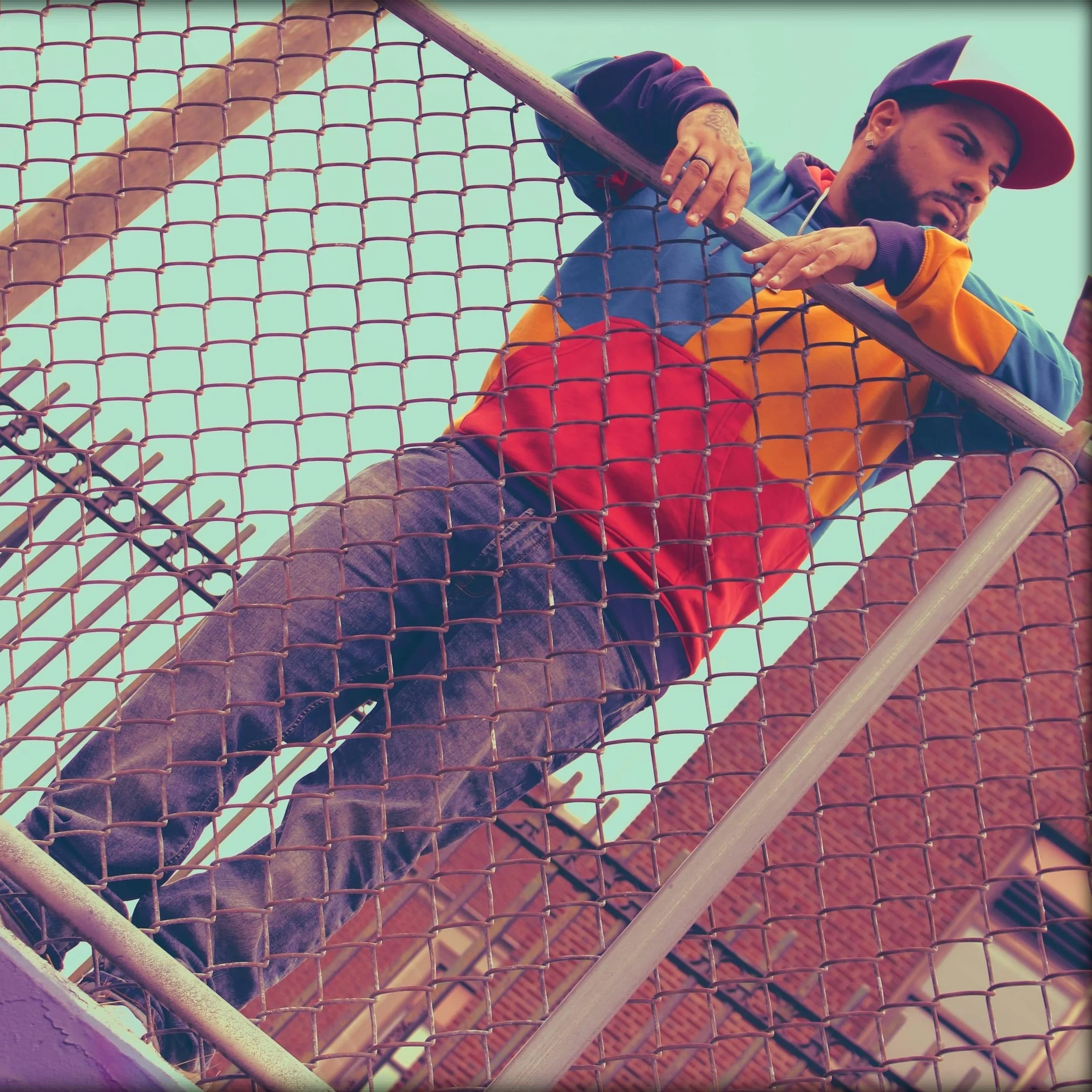 A person with a tattooed hand resting on a chain-link fence, wearing a colorful hoodie and a cap, with a brick building in the background.
