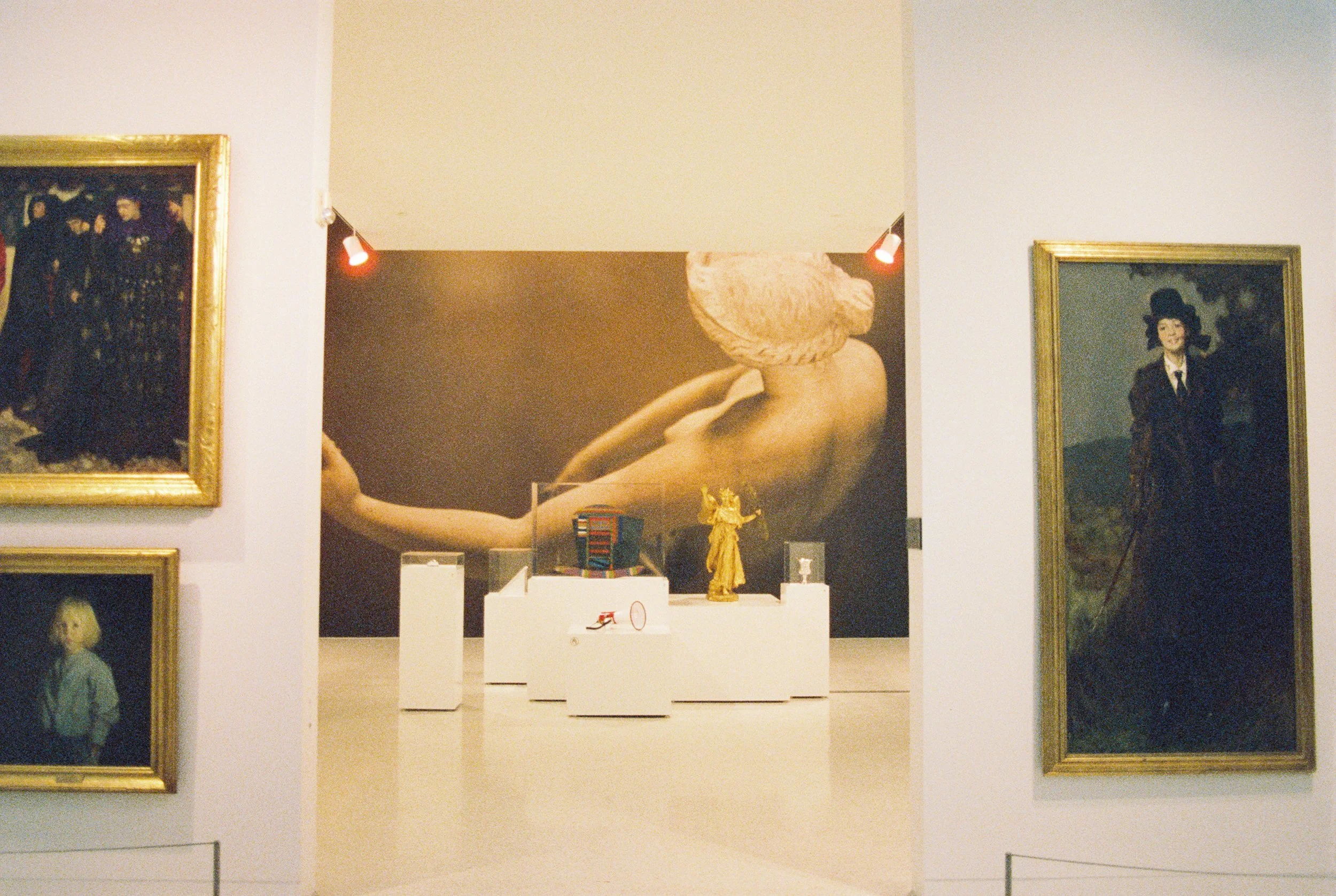 Art gallery with framed paintings on white walls, centered behind white display pedestals with sculptures and objects, and a large background mural of a reclining nude woman with a cloth wrapped around her head.