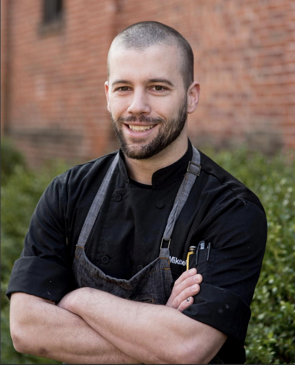 A smiling man with a beard and short hair, dressed in a black chef's coat and apron, standing outdoors with a brick wall and greenery in the background.