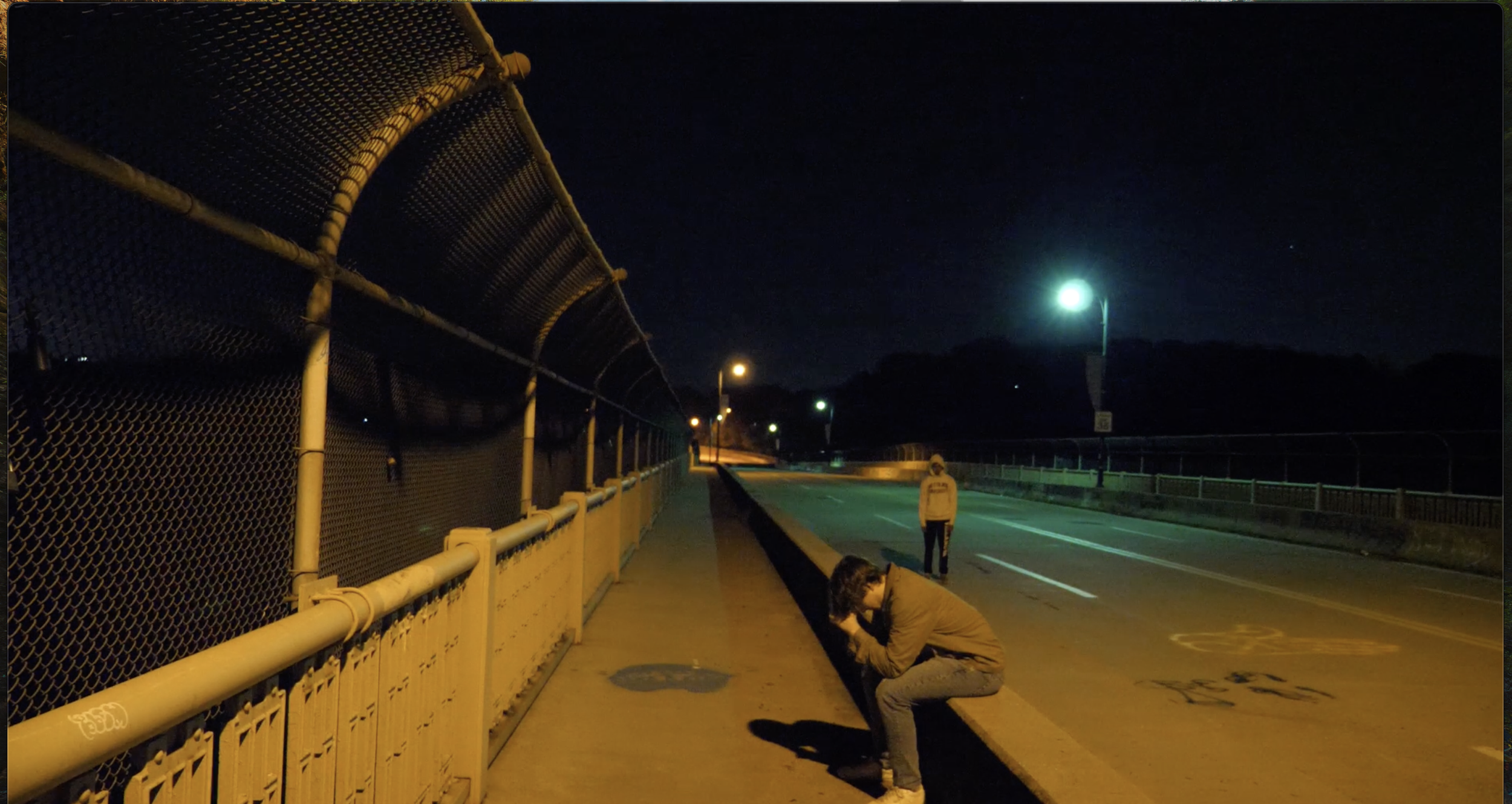 Nighttime scene on a quiet street with two people; one is squatting near the sidewalk taking a photo or looking closely at something, and the other is standing further down the road. Streetlights illuminate the scene along a sidewalk with a chain-link fence on one side and empty road markings on the other.