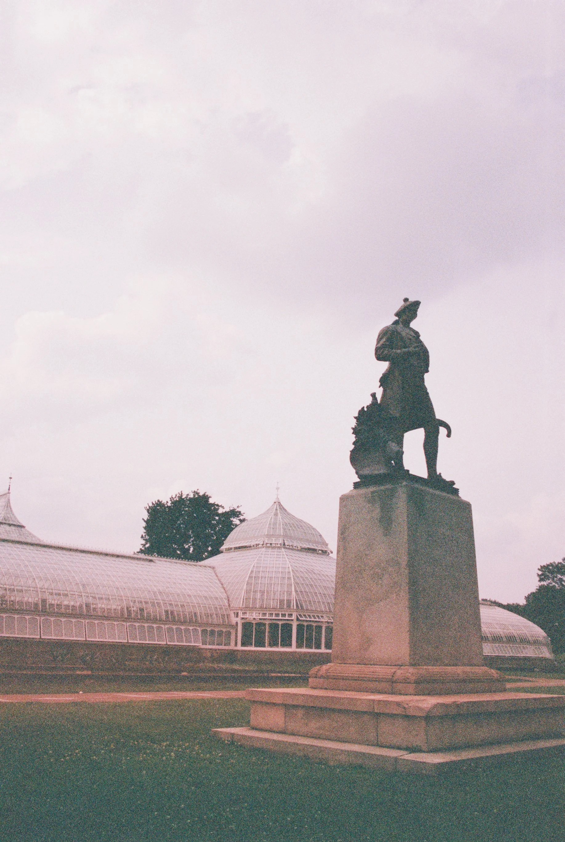 A statue of a man standing on a pedestal, with a greenhouse or conservatory building in the background under an overcast sky.