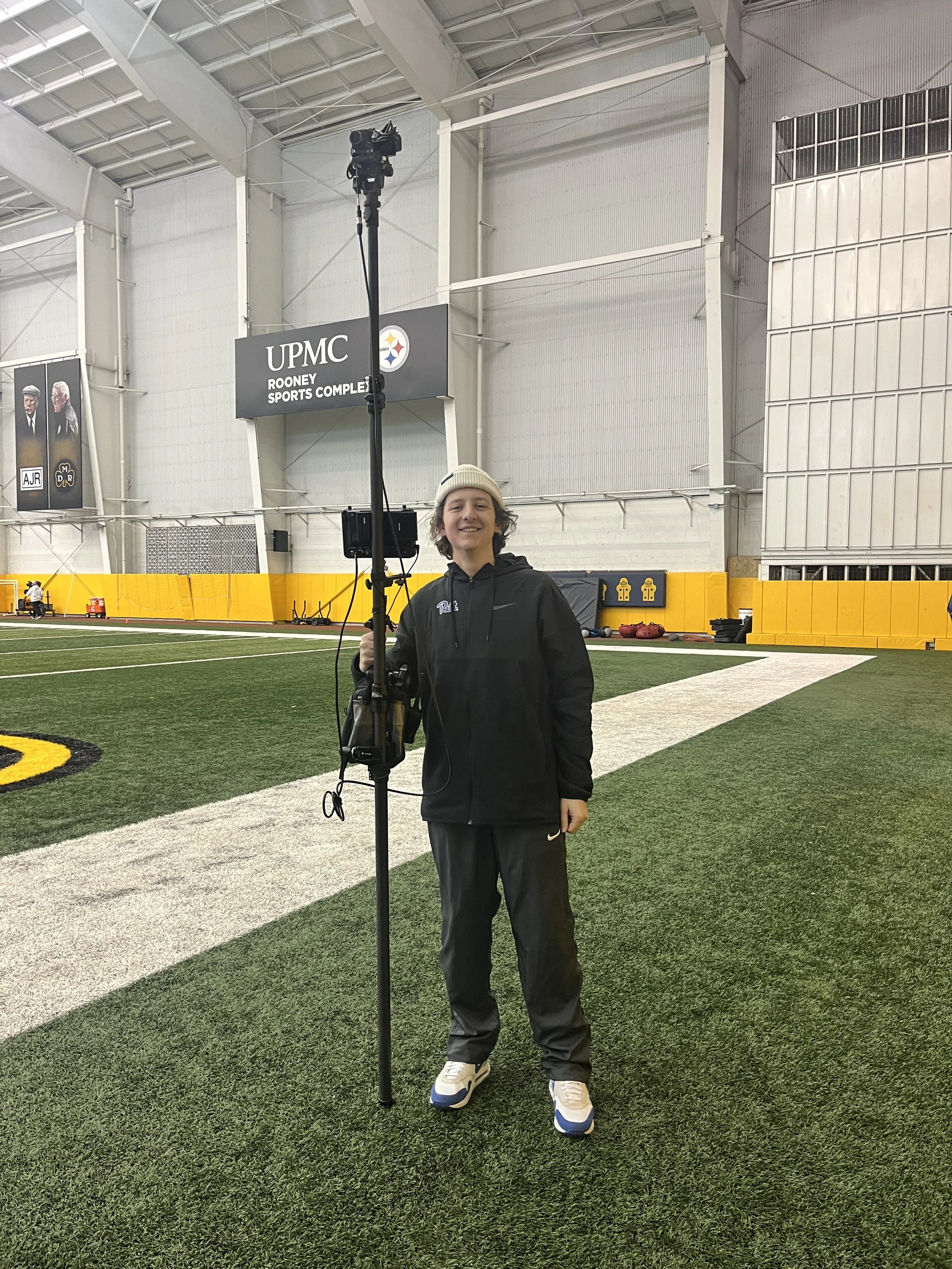 A person standing on an indoor football field holding a camera on a tall tripod, smiling at the camera, inside UPMC Rooney Sports Complex.