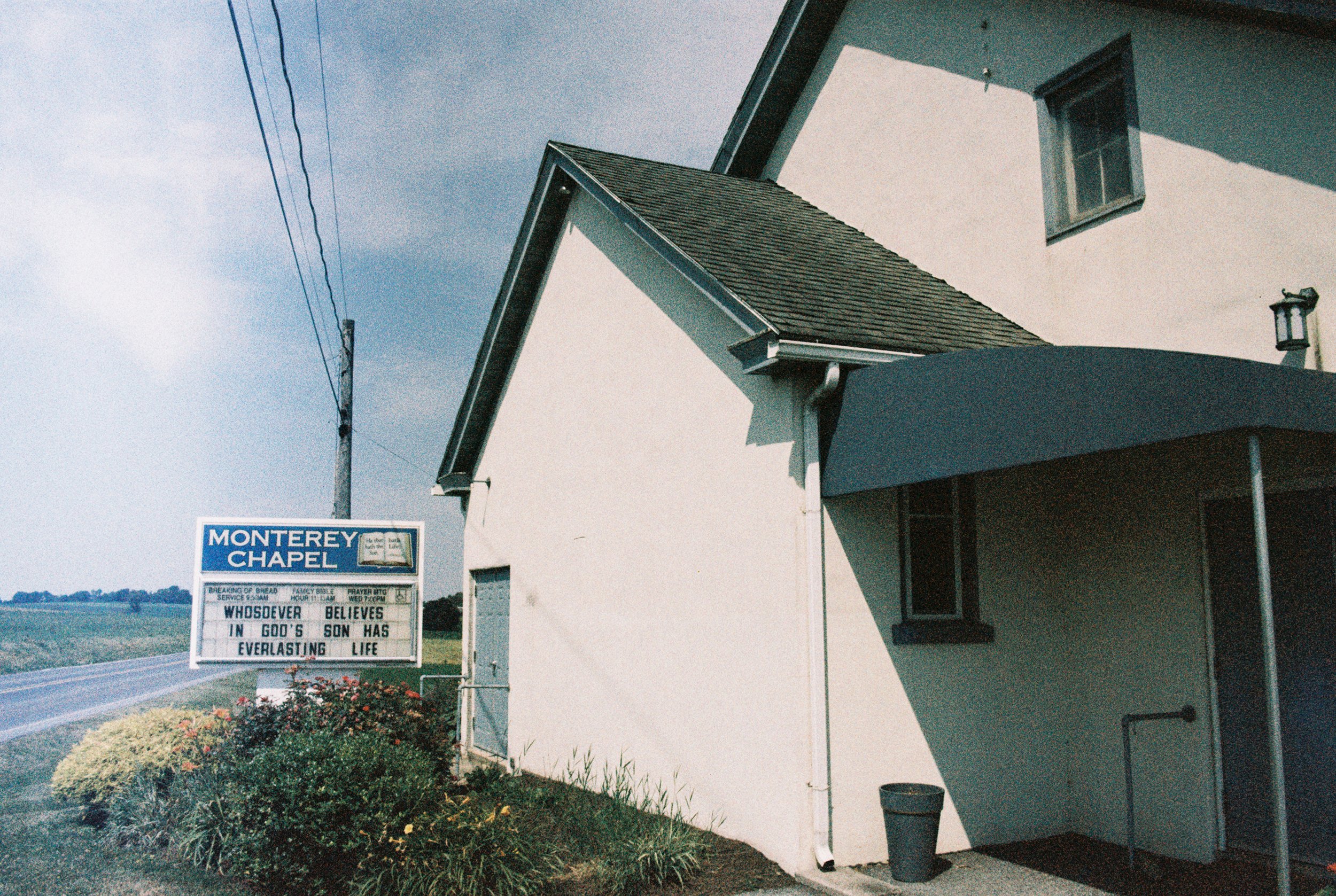 A church building with a sign that reads Monterey Chapel, located along a rural roadside with bushes and flowers in front, under a partly cloudy sky.