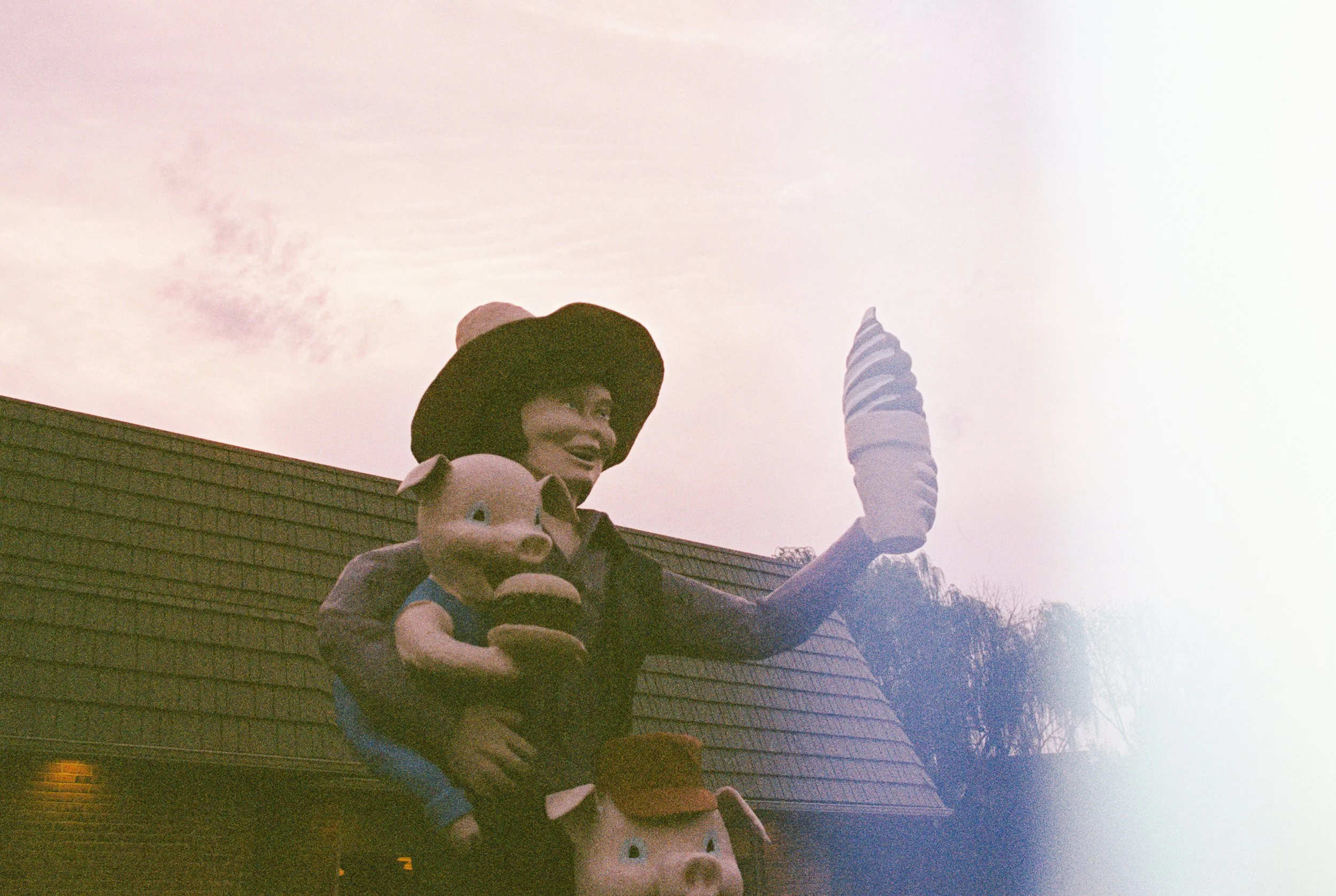 A statue wearing a wide-brimmed hat and dark clothing holding two pig-shaped toys. One toy pig is on their shoulder, and the other is held in front of them. The background shows a building with a sloped roof and a sky with light clouds.