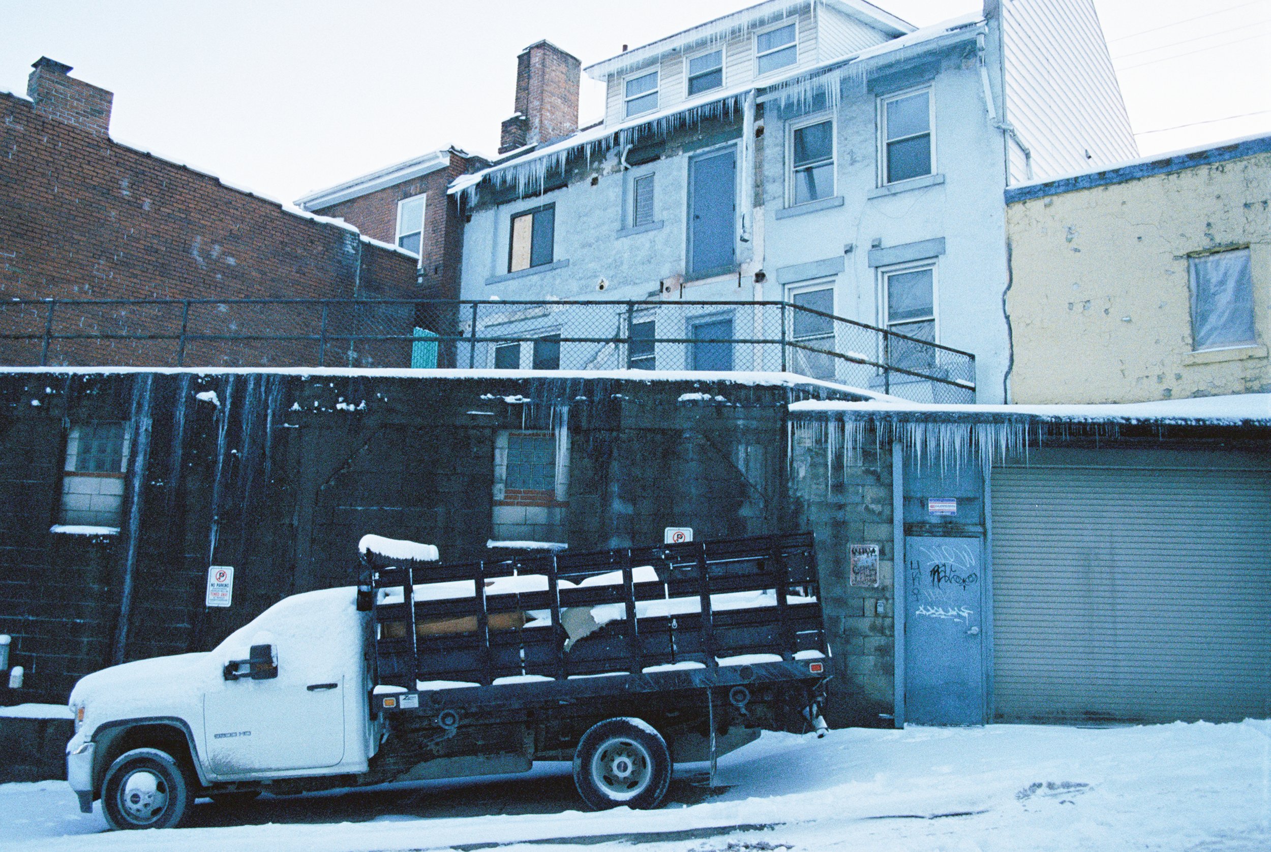 A snow-covered urban alleyway with a white flatbed truck parked in front of a building with graffiti and graffiti-covered garage doors, icicles hanging from the roof, and multi-story houses above.