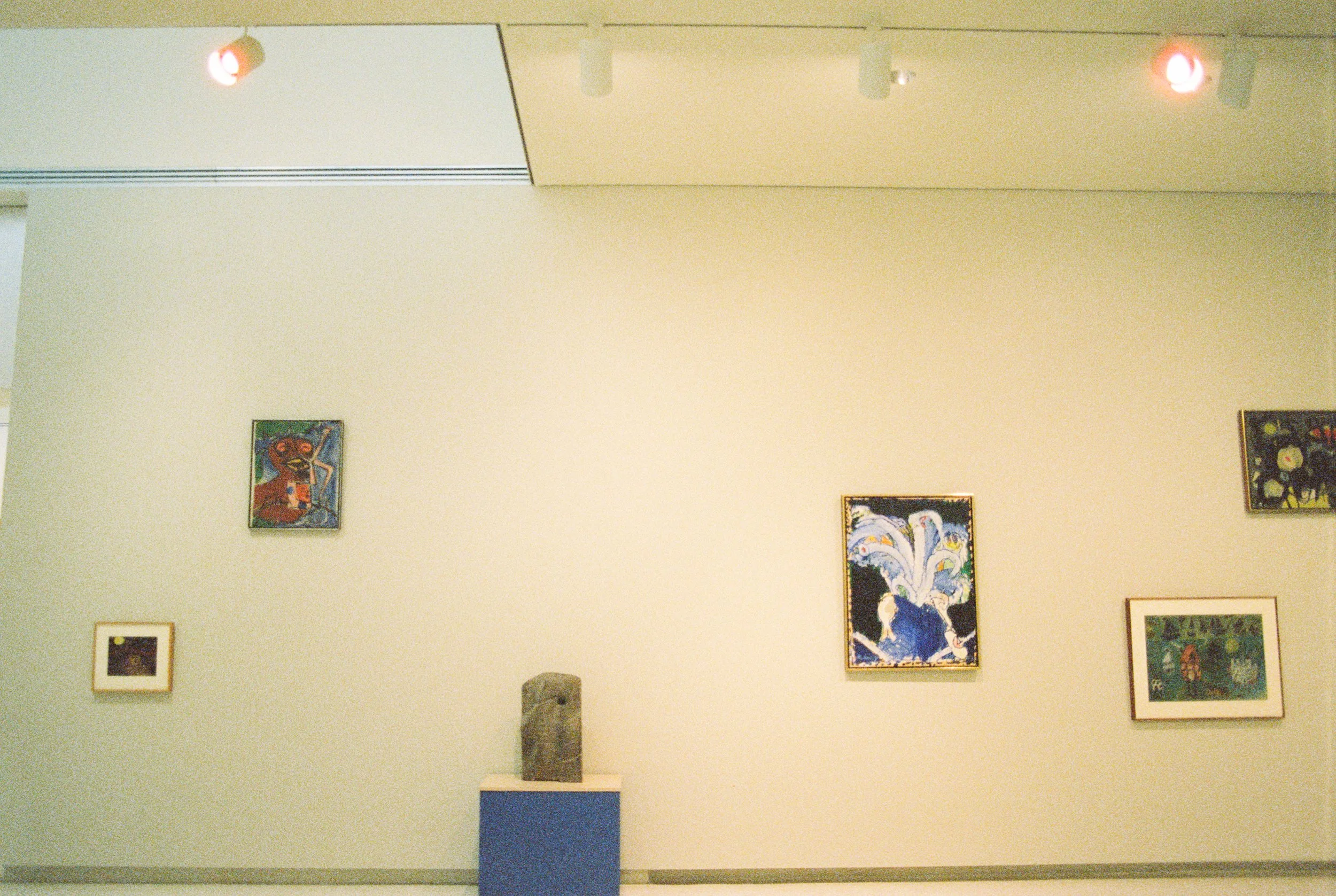 An art gallery wall displaying various framed paintings and a sculpture on a blue pedestal.