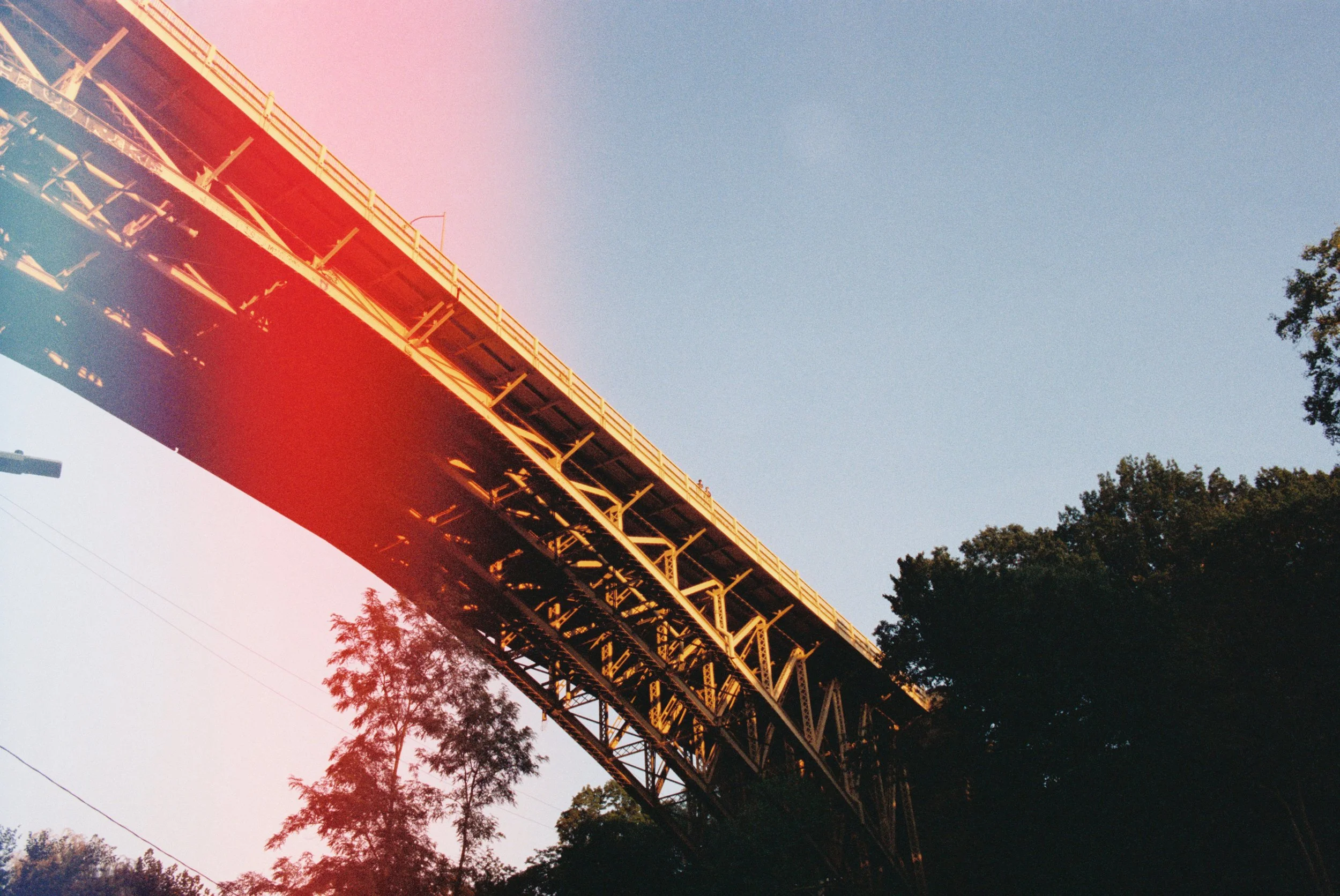 A large bridge with a metal framework seen from below against a clear blue sky, with trees at the bottom of the image.