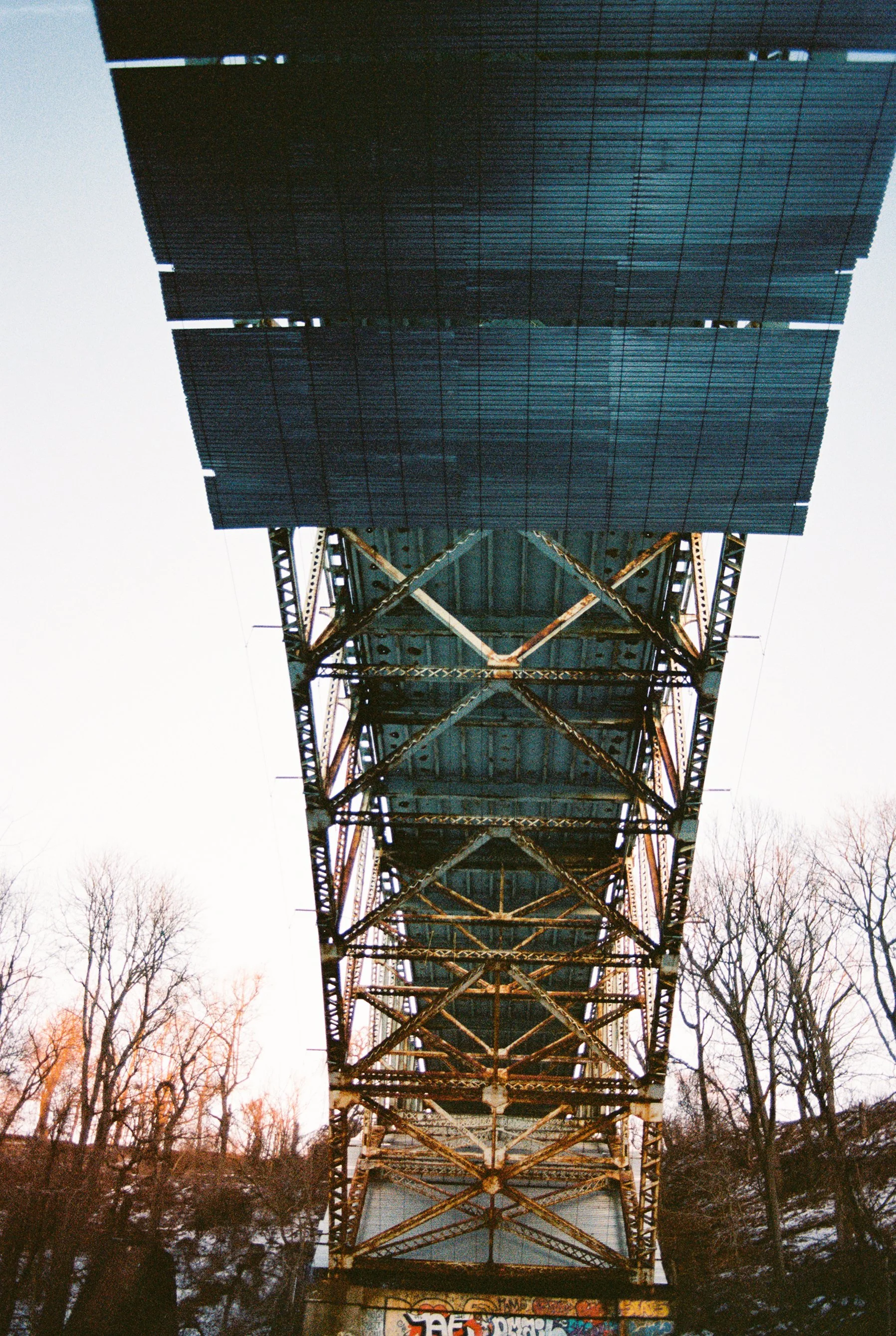 View of the underside of a bridge with a metal structure and solar panels on top, alongside graffiti art at the bottom, with trees in the background.