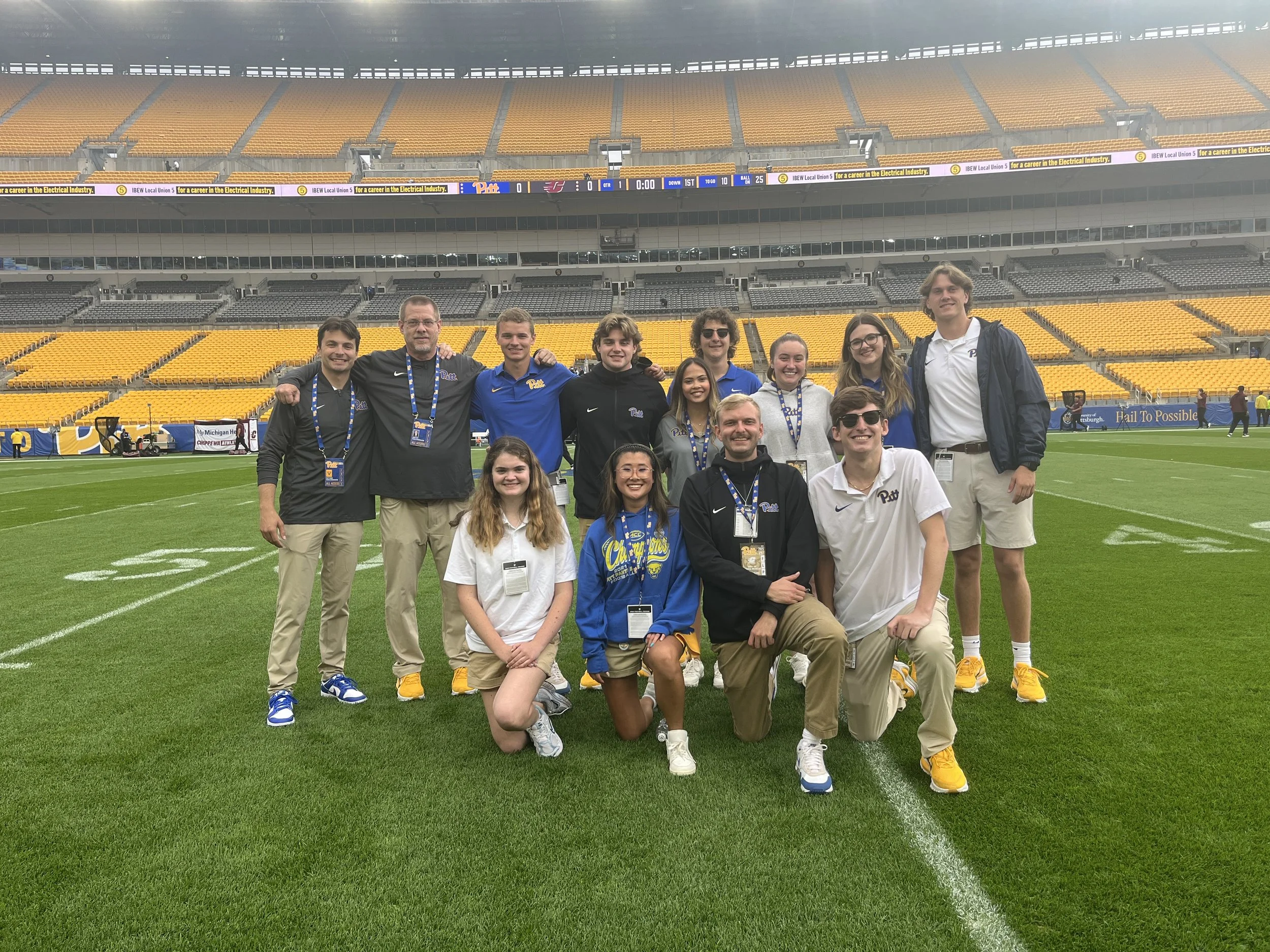 Group of young people and adults standing on a football field inside a stadium, smiling for the photo, some wearing University of Pittsburgh apparel.