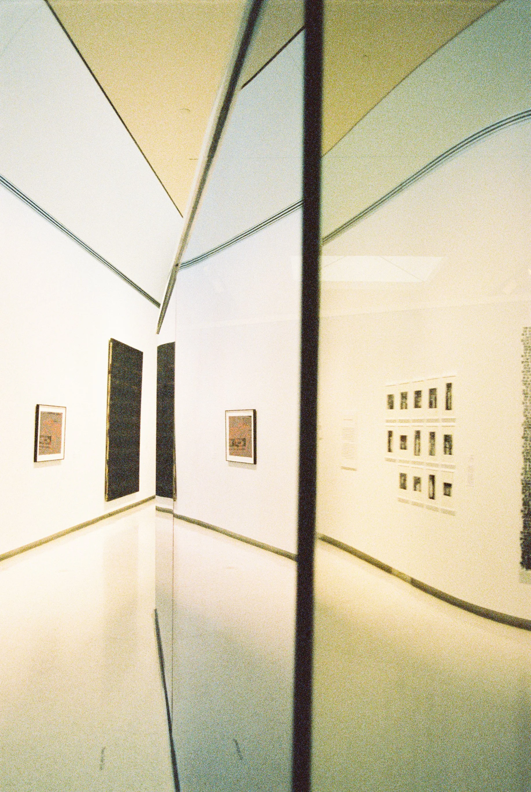 A mirror in an art gallery reflects a white wall with framed artwork and an adjacent wall with a display of smaller pictures.