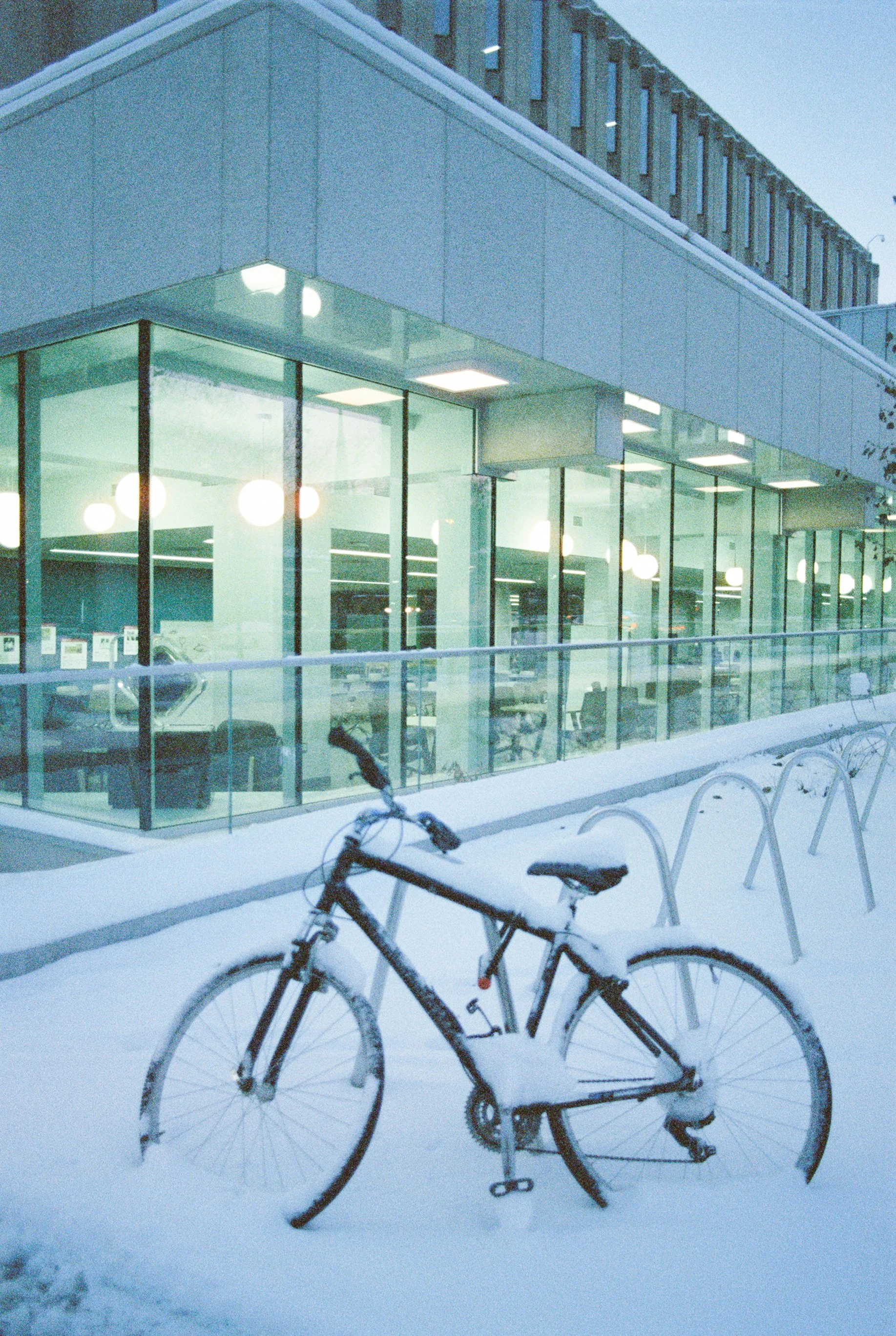 A bicycle covered in snow outside a modern glass building at dusk.