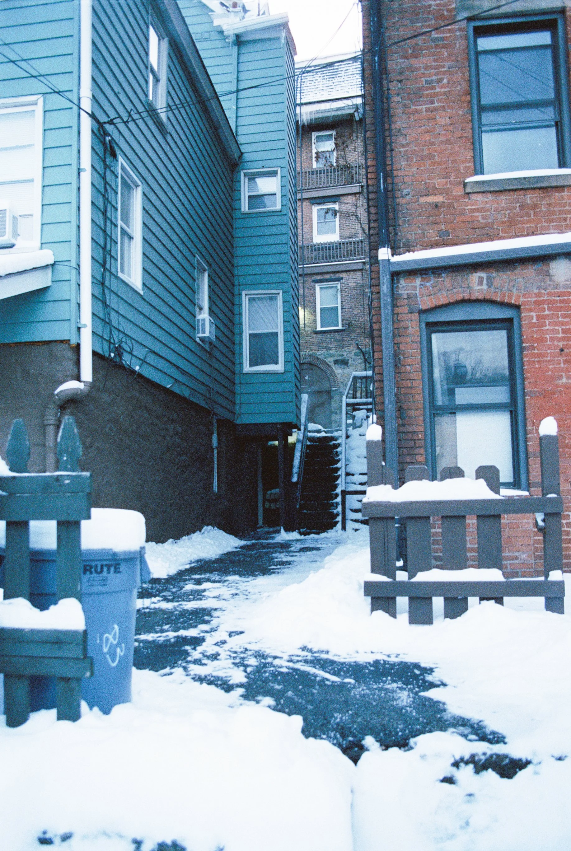 Snow-covered alleyway between two buildings, one painted blue and the other brick, with a stairway leading up between the buildings and snow on the ground.