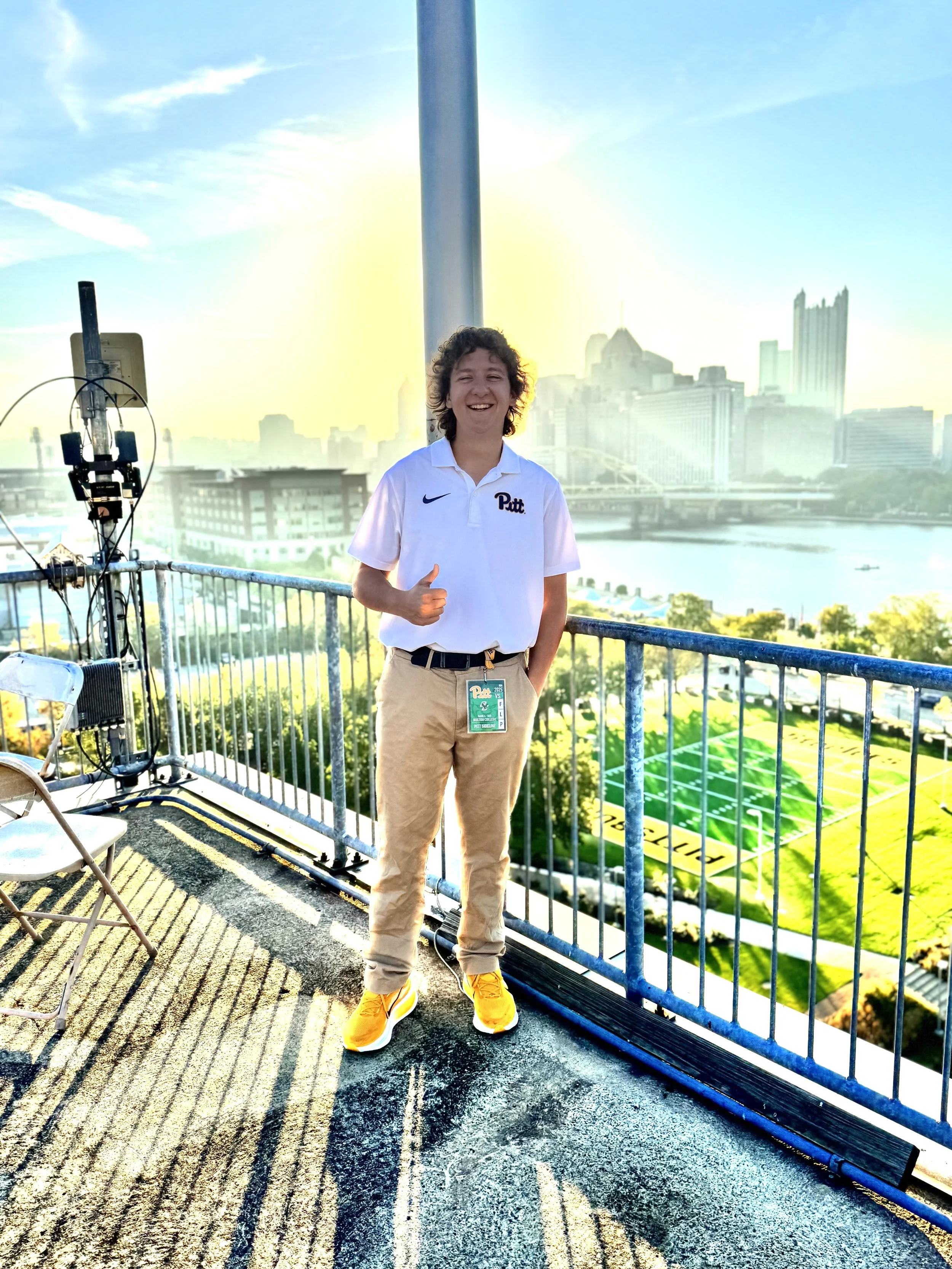 A young man standing on a rooftop balcony during sunset, wearing a white Pitt polo shirt, khaki pants, and yellow sneakers, with a city skyline and river in the background.