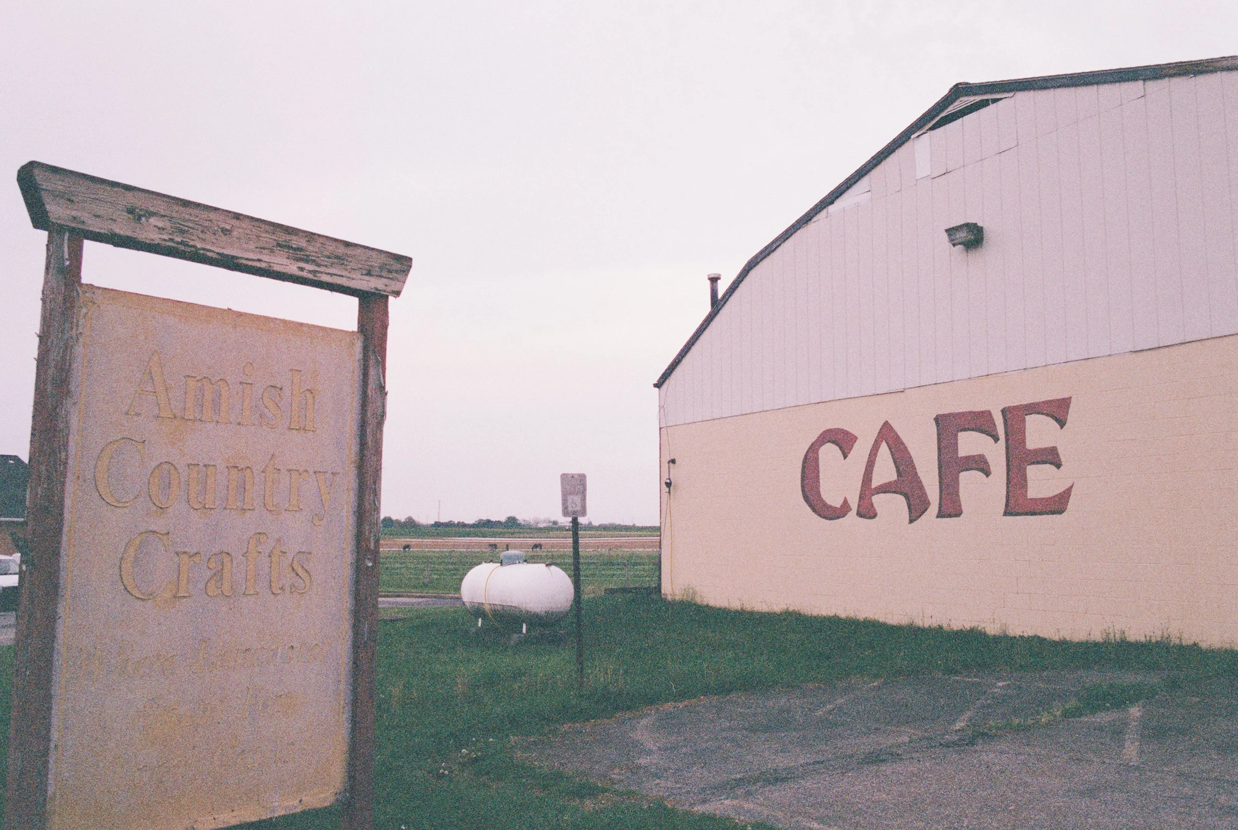 A weathered sign reading 'Amish Country Crafts' beside a large beige building with pink and red signs that says 'CAFE' in a rural area with a propane tank in the background.