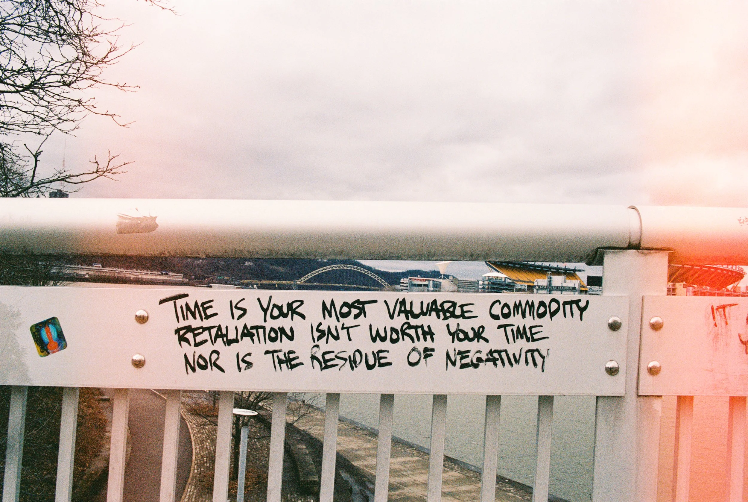 A bridge railing with a handwritten quote that says, 'Time is your most valuable commodity. Retrospection isn't worth your time nor is the respite of negativity.' In the background, a river, trees, and a cityscape with a Yellow stadium and a bridge a