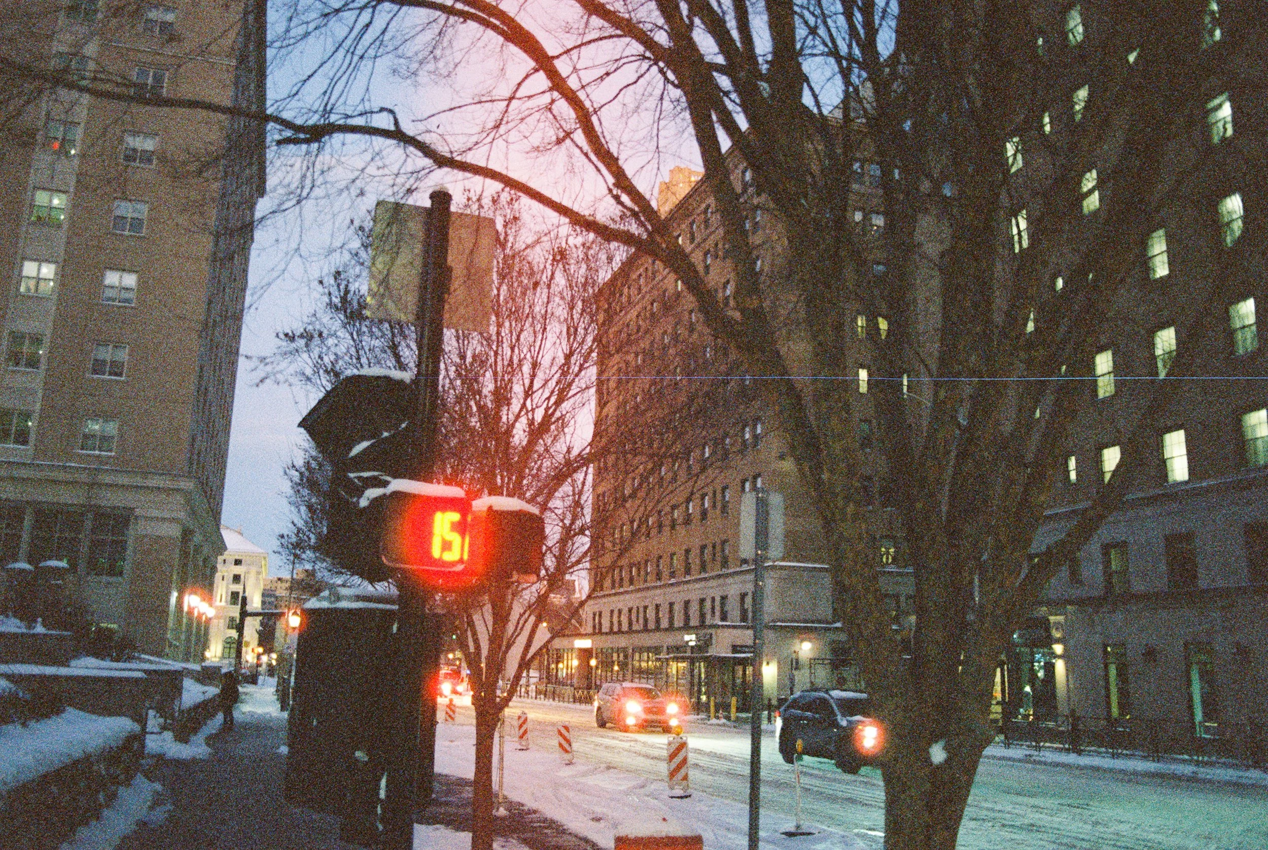 A snowy city street scene at dusk with tall buildings, leafless trees, and cars driving. A traffic signal displays the number 15 in red.