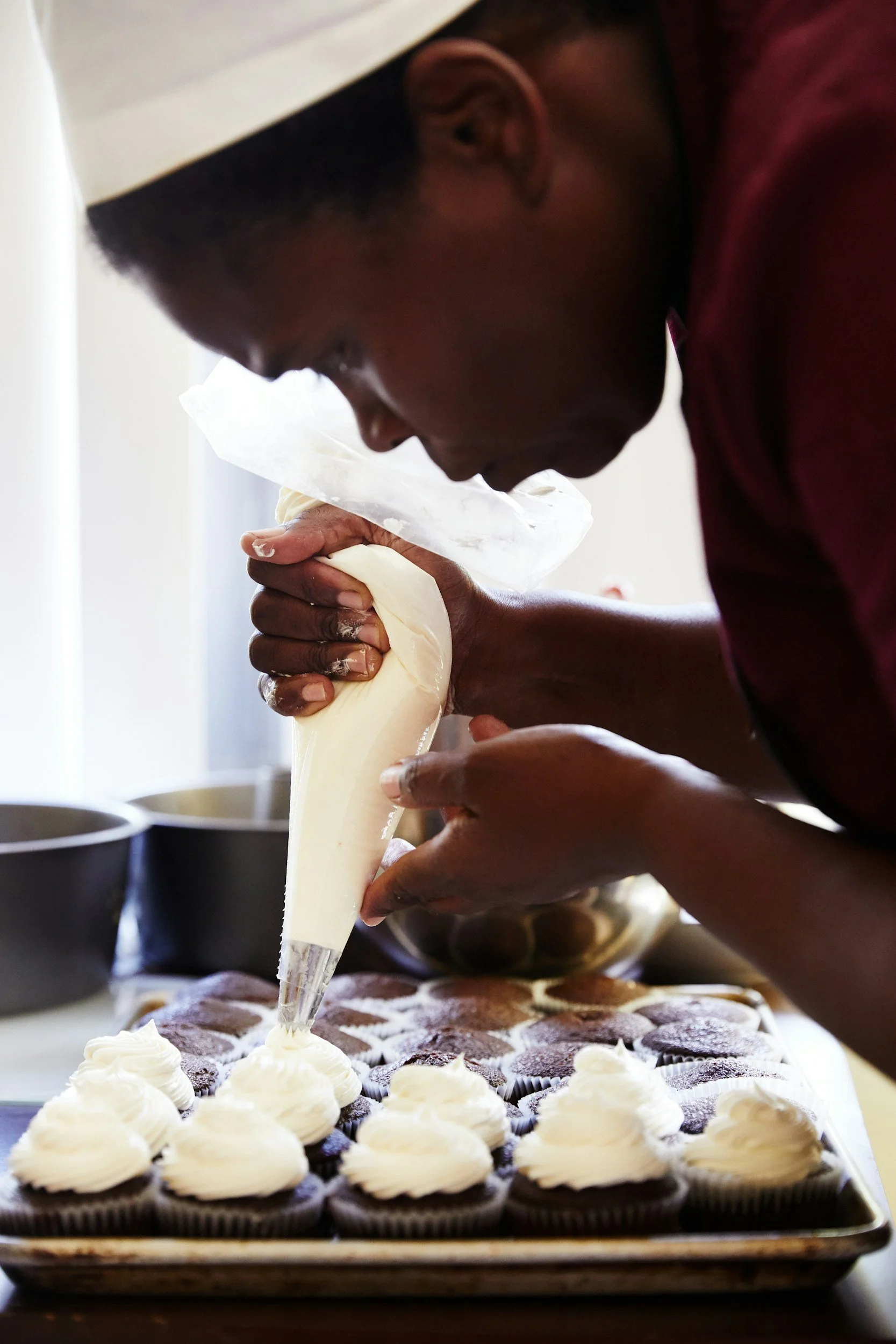A person in a chef's hat decorators cupcakes with white frosting from a piping bag.