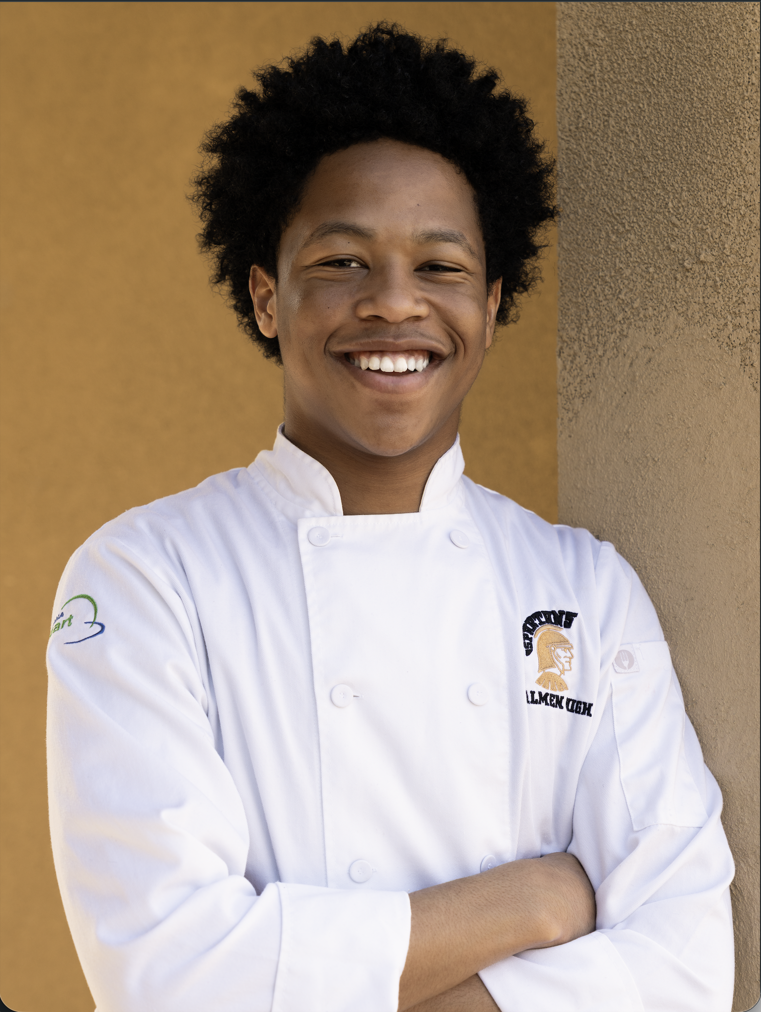 Portrait of a young African American man with curly hair, smiling, wearing a white chef's coat, standing against a beige and brown textured wall.