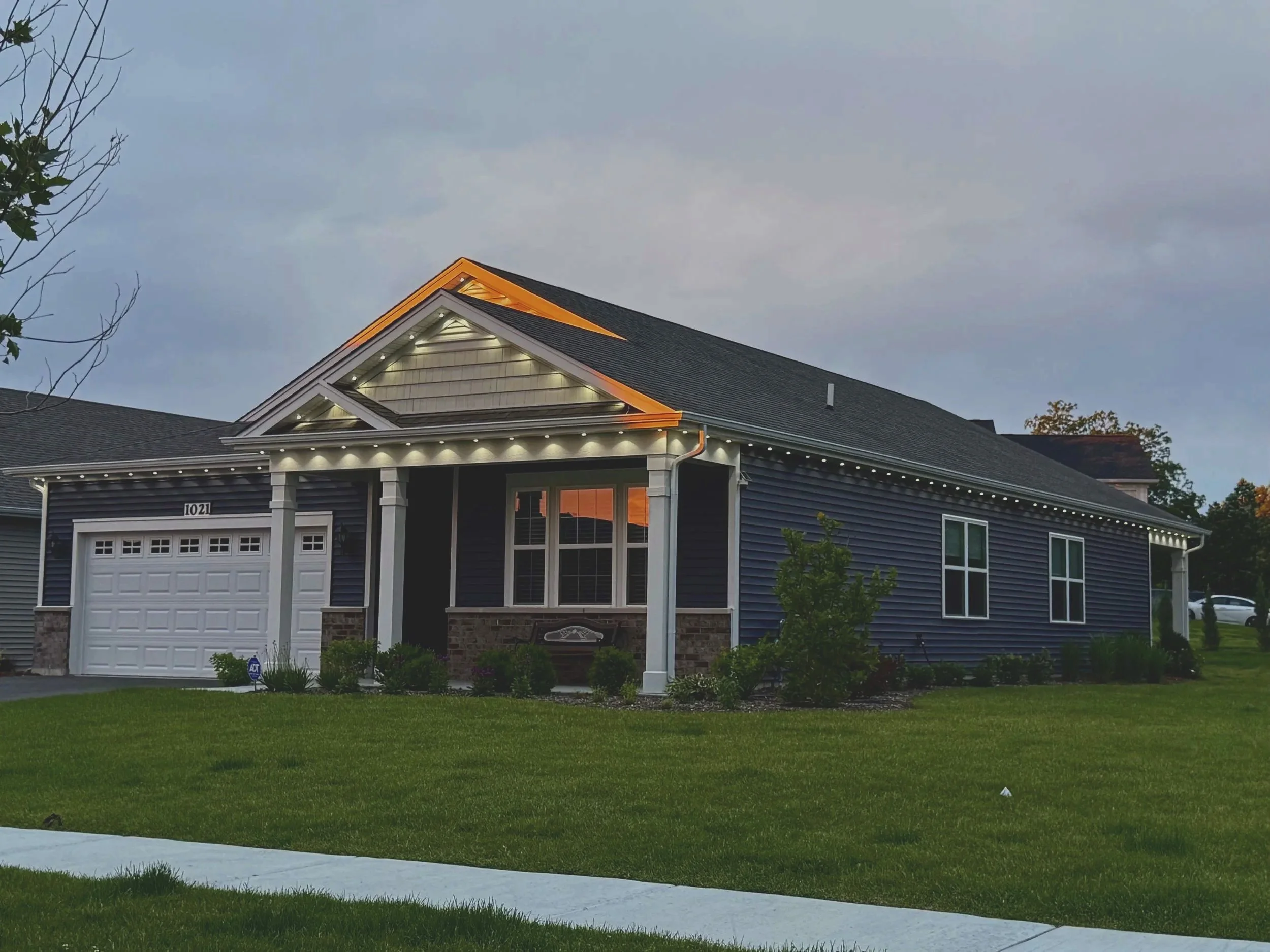 Front view of a modern house with a dark gray exterior, white trim, a two-car garage, and a front porch, taken during dusk with the sky partly cloudy. The house has outdoor lighting, a manicured lawn, and some trees and shrubs around.