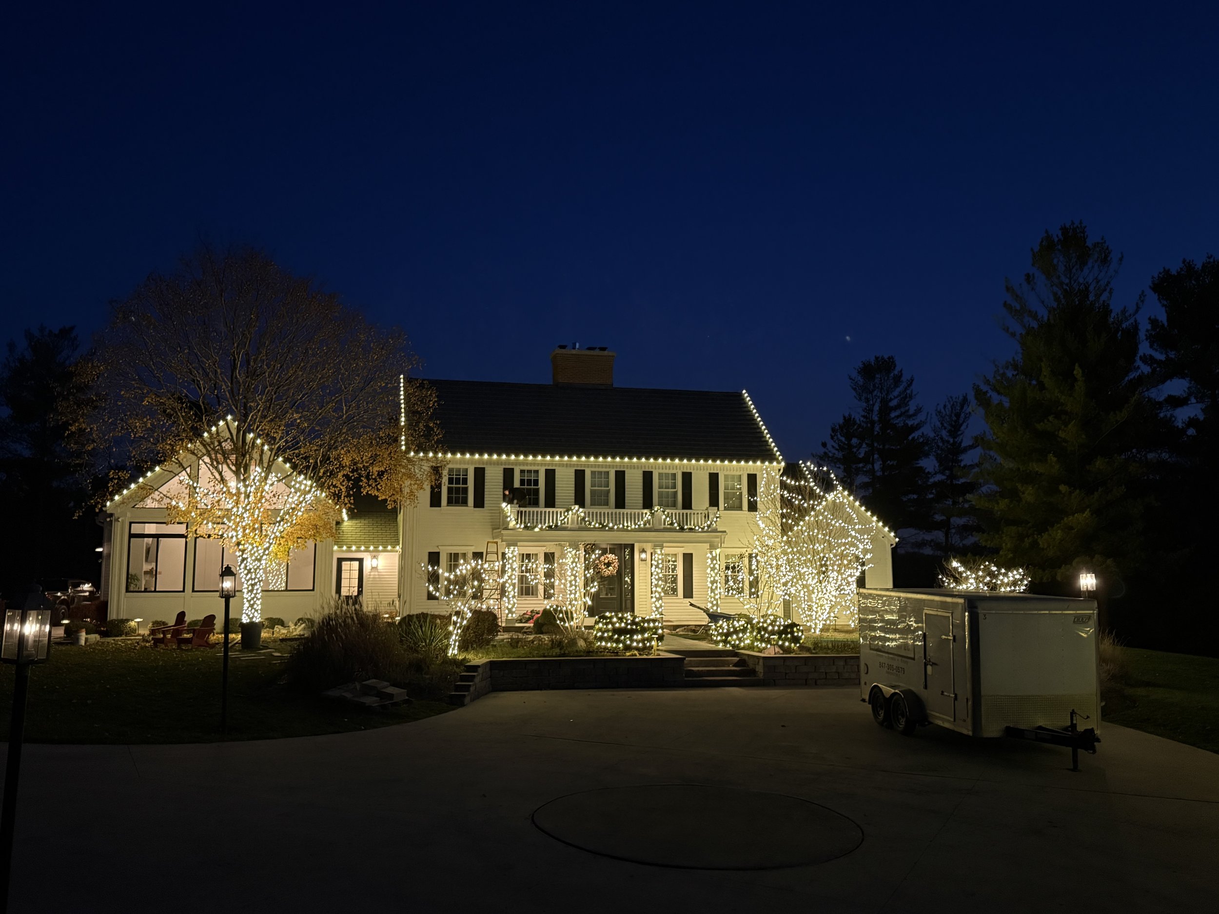 Large house decorated with white string lights outlining the roof, windows, and trees in front. The house has a porch with a wreath, and there are trees and shrubs decorated with lights. The night sky is dark blue with a few visible stars.