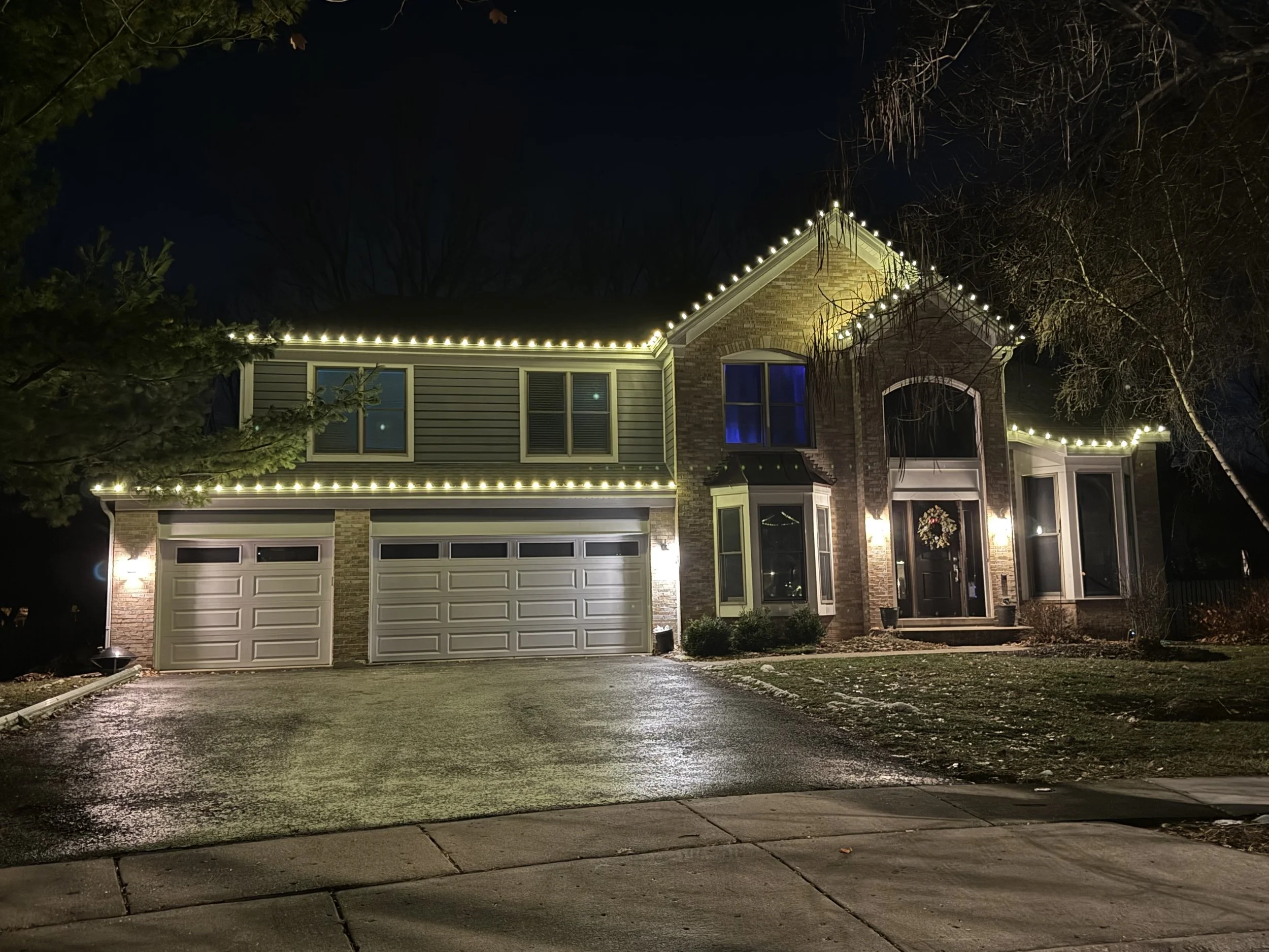 A two-story house decorated with string lights along the roofline at night. The house features a brick facade and multiple windows, with a dark sky in the background. The driveway and sidewalk are visible in the foreground, with some patches of grass
