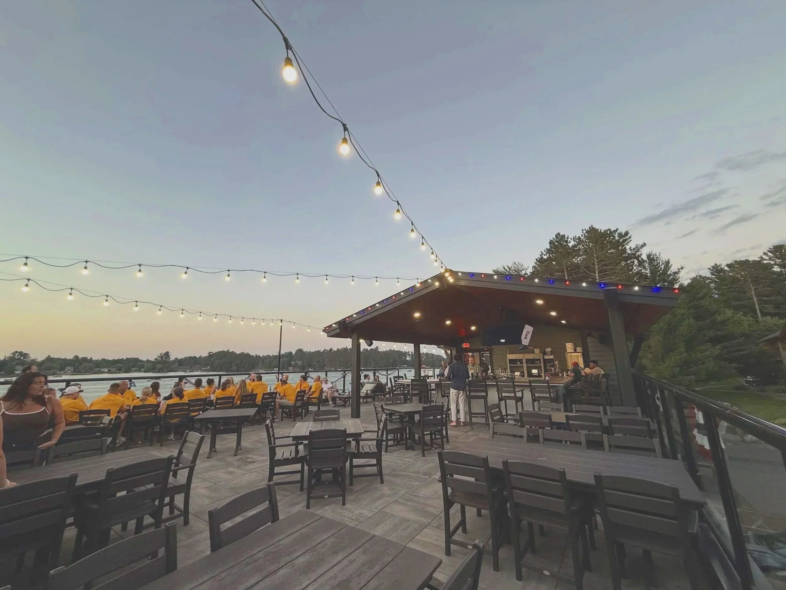 Outdoor patio at dusk with string lights overhead and a gathering of people, some wearing yellow shirts, near a body of water and surrounded by trees.