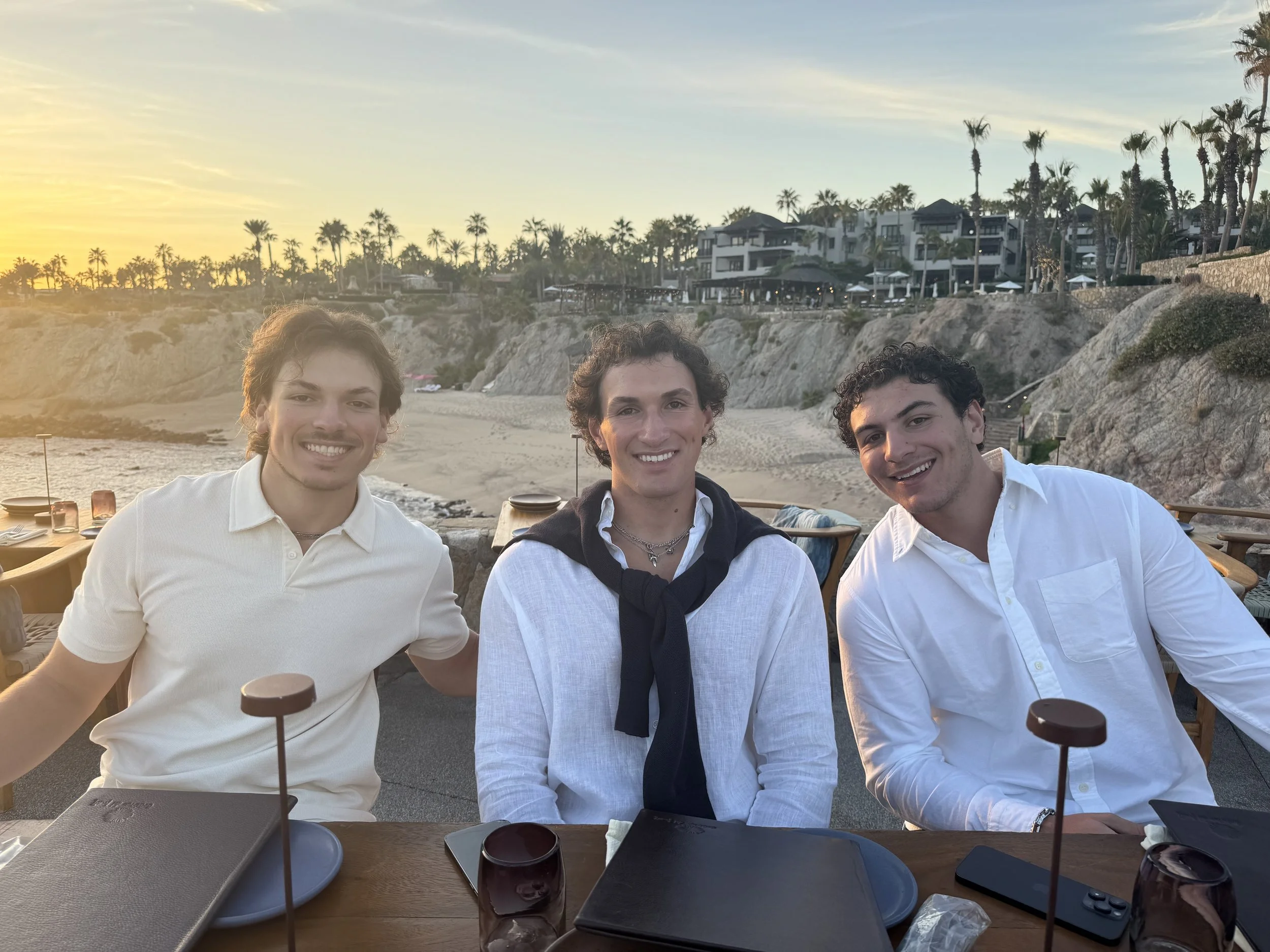 Three young men sitting at a restaurant table outdoors during sunset on a beach with cliffs and houses in the background.
