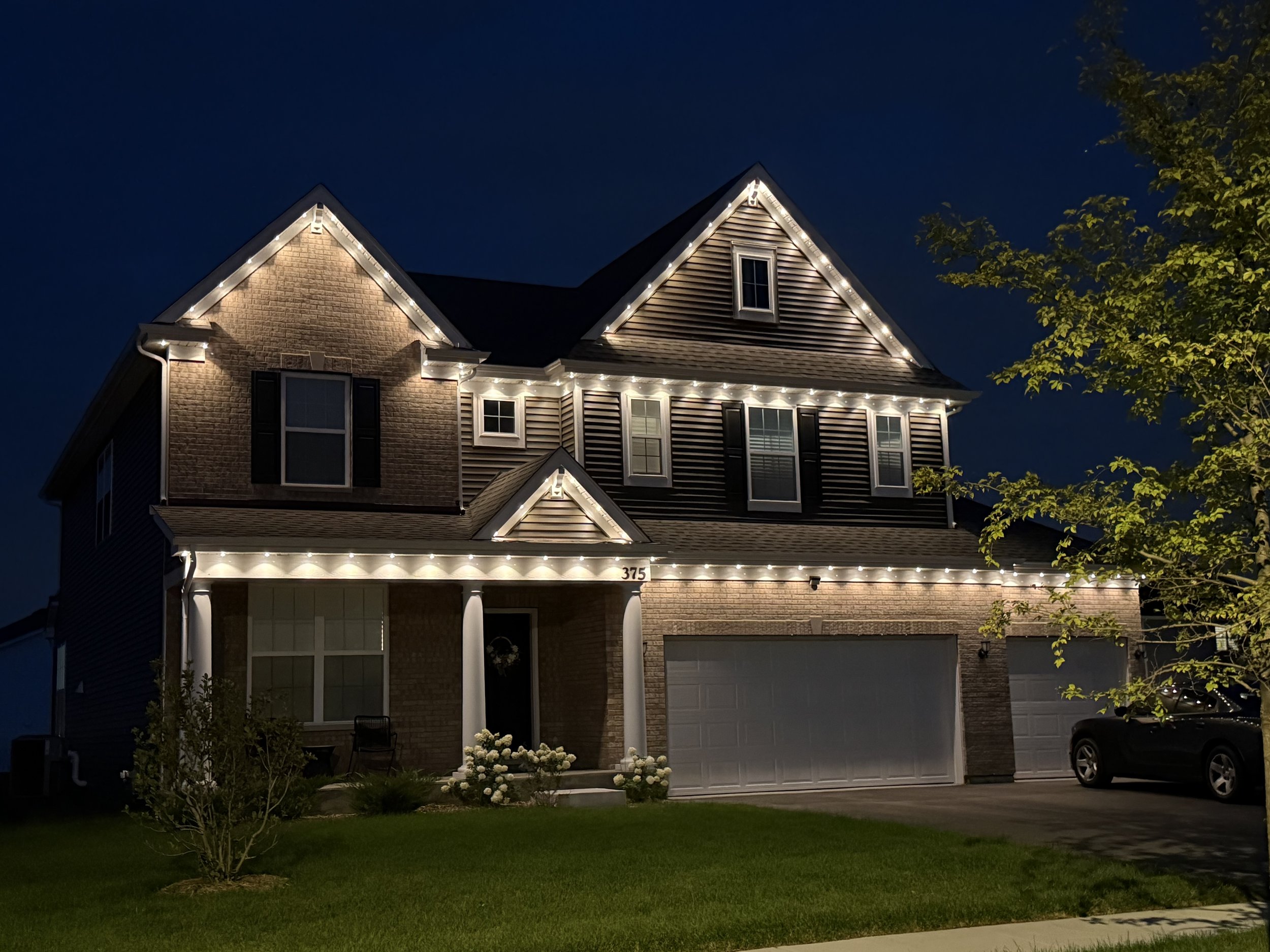 A two-story house decorated with white string lights along the roofline and around the windows, with a small front yard, bushes, and a tree, during early evening or night.