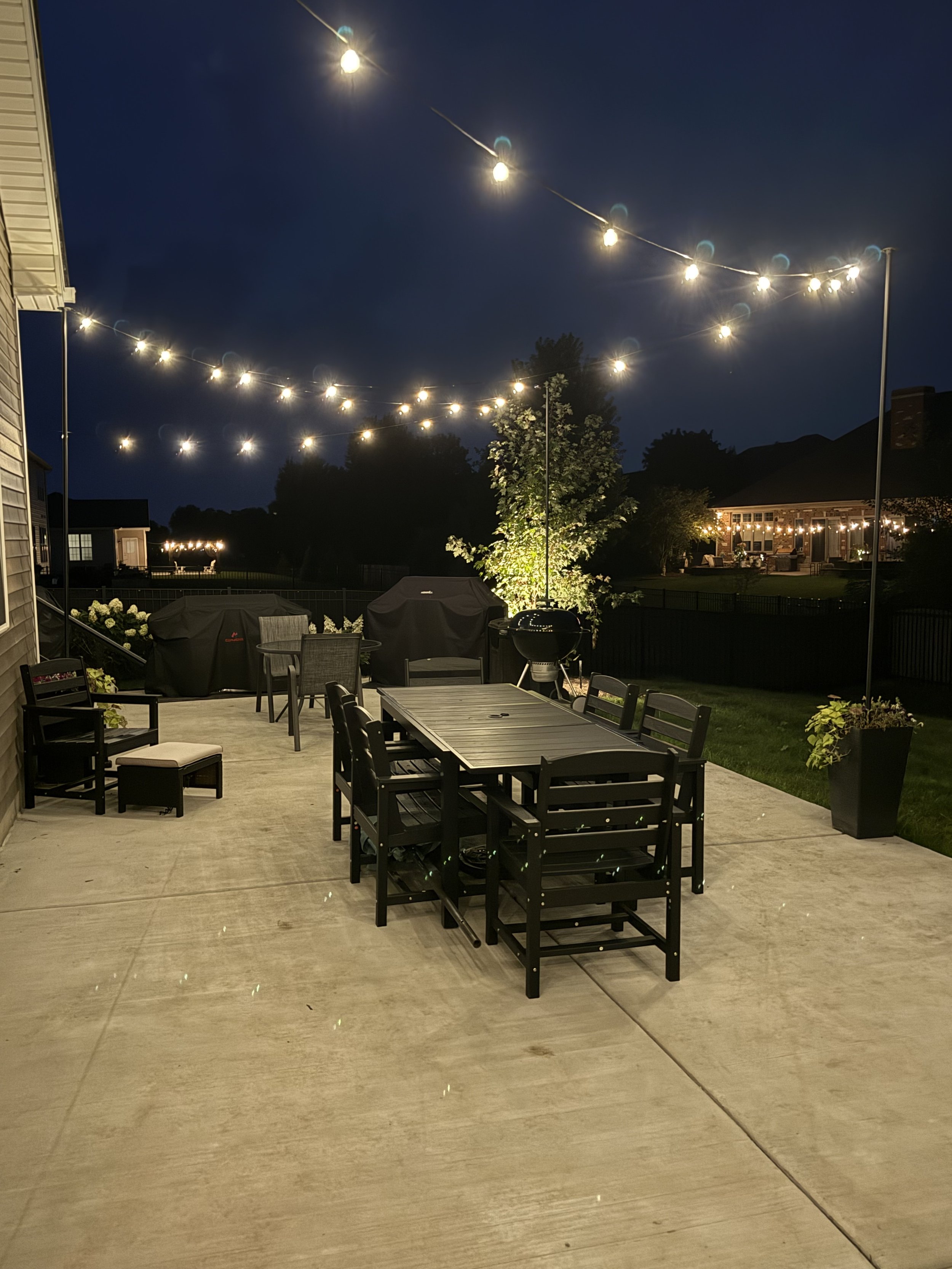 An outdoor patio at night with string lights hanging above, a grill, patio furniture, and a yard with trees and a house in the background.
