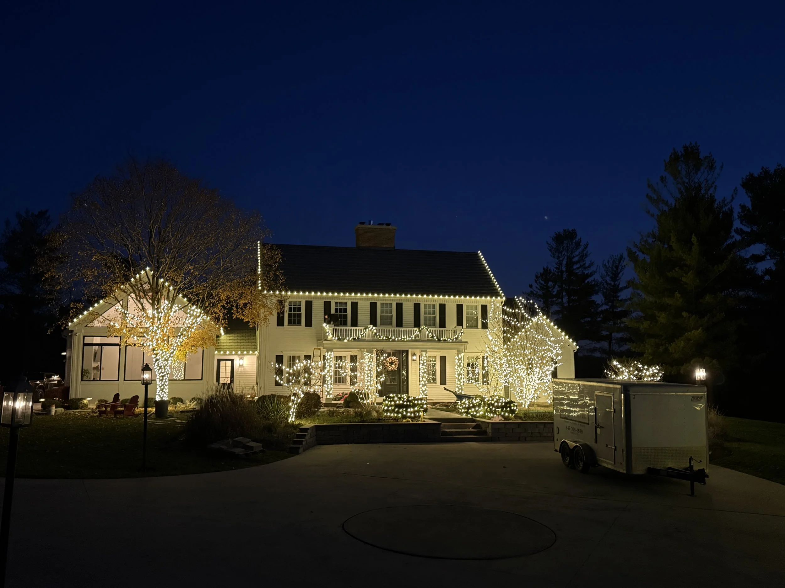 A house decorated with white string lights outlining the roof, porch, and trees, with a dark blue night sky and a few visible stars and a planet.